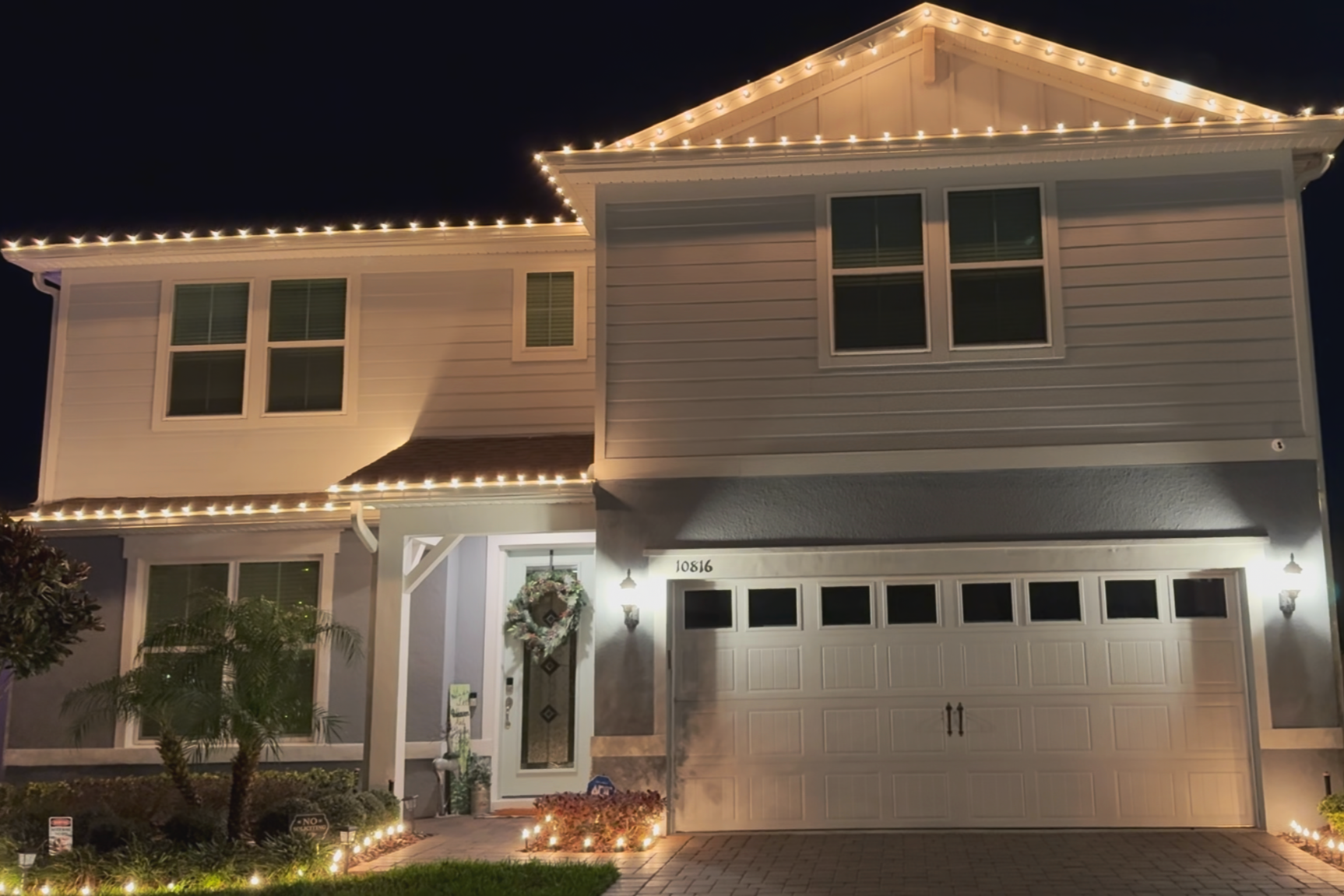 Person standing on a ladder hanging red holiday lights on a wooden garage exterior.