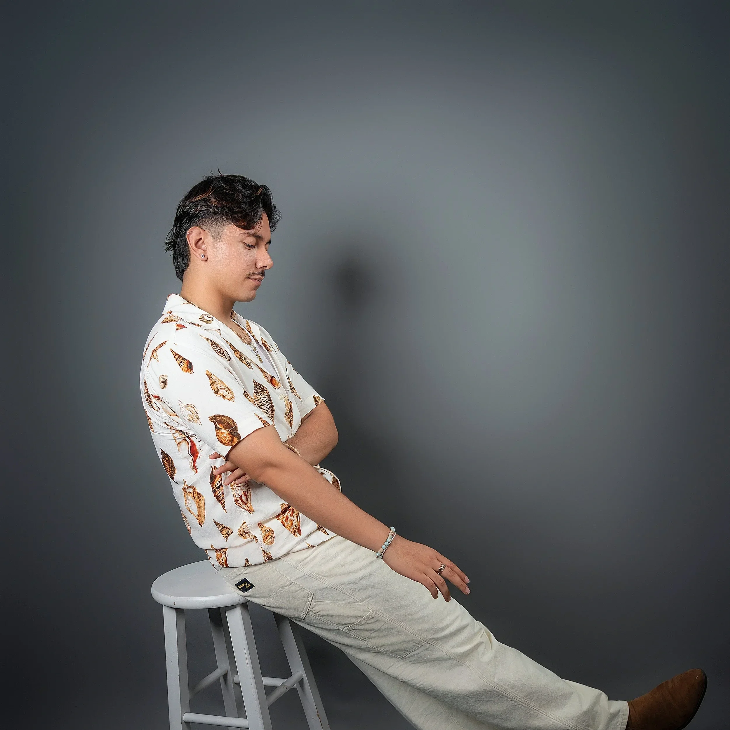 A young man with dark hair and a mustache, seated sideways on a white stool against a dark gray background, wearing a white short-sleeved shirt with a seashell pattern, cream-colored pants, and brown shoes, with his arms crossed and eyes closed.