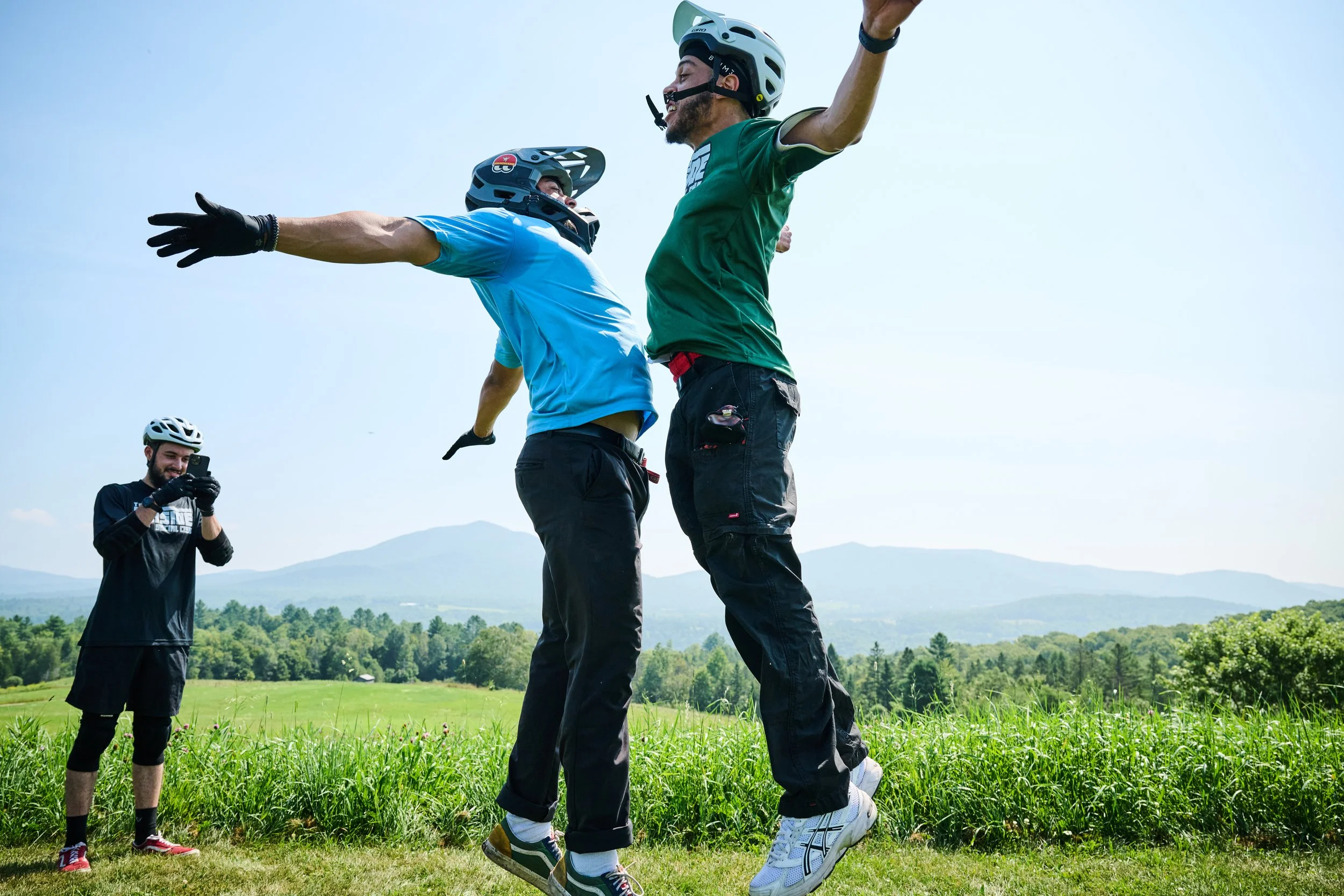 Two men wearing mountain biking helmets and outdoor clothing are mid-air jumping and celebrating in a grassy field with mountains in the background. A younger person in the background is taking a photo of them. East Burke VT