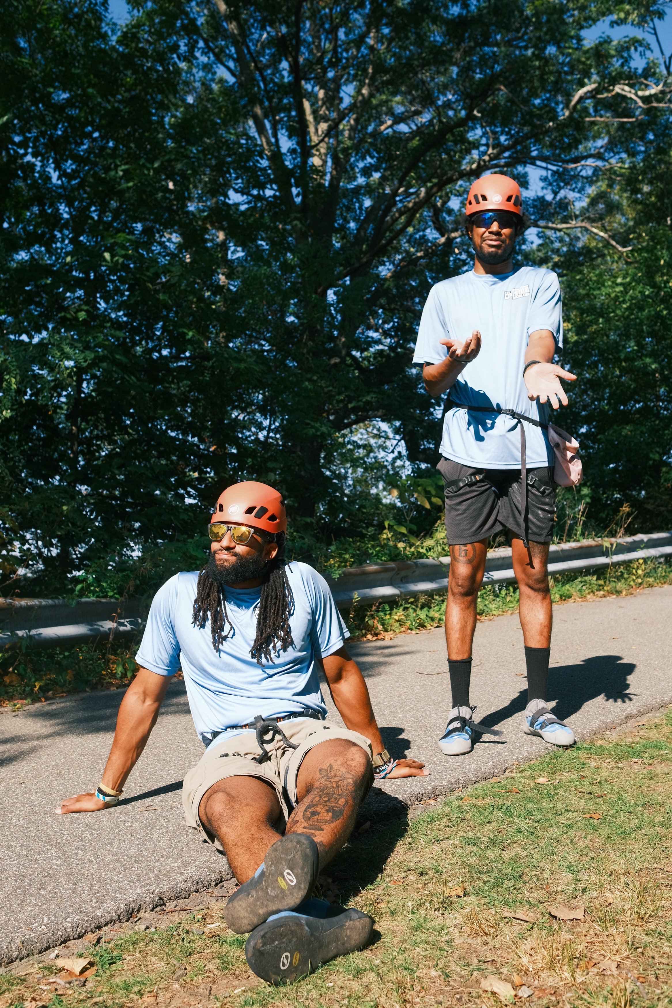 Two men in blue t-shirts and rock climbing helmets and rock climbing shoes, one sitting on the ground and the other standing, outdoors on a paved path surrounded by trees.