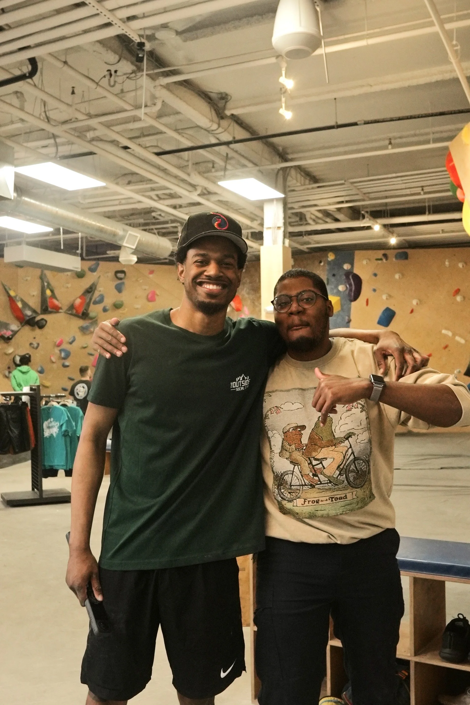 Two men standing together at an indoor rock climbing gym, smiling, with one man wearing a green t-shirt and the other a beige t-shirt with a graphic of two bears riding a bicycle.