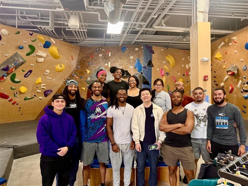 Group of diverse people, including men and women, smiling and posing for a photo inside a rock climbing gym with colorful climbing holds on the walls in the background.