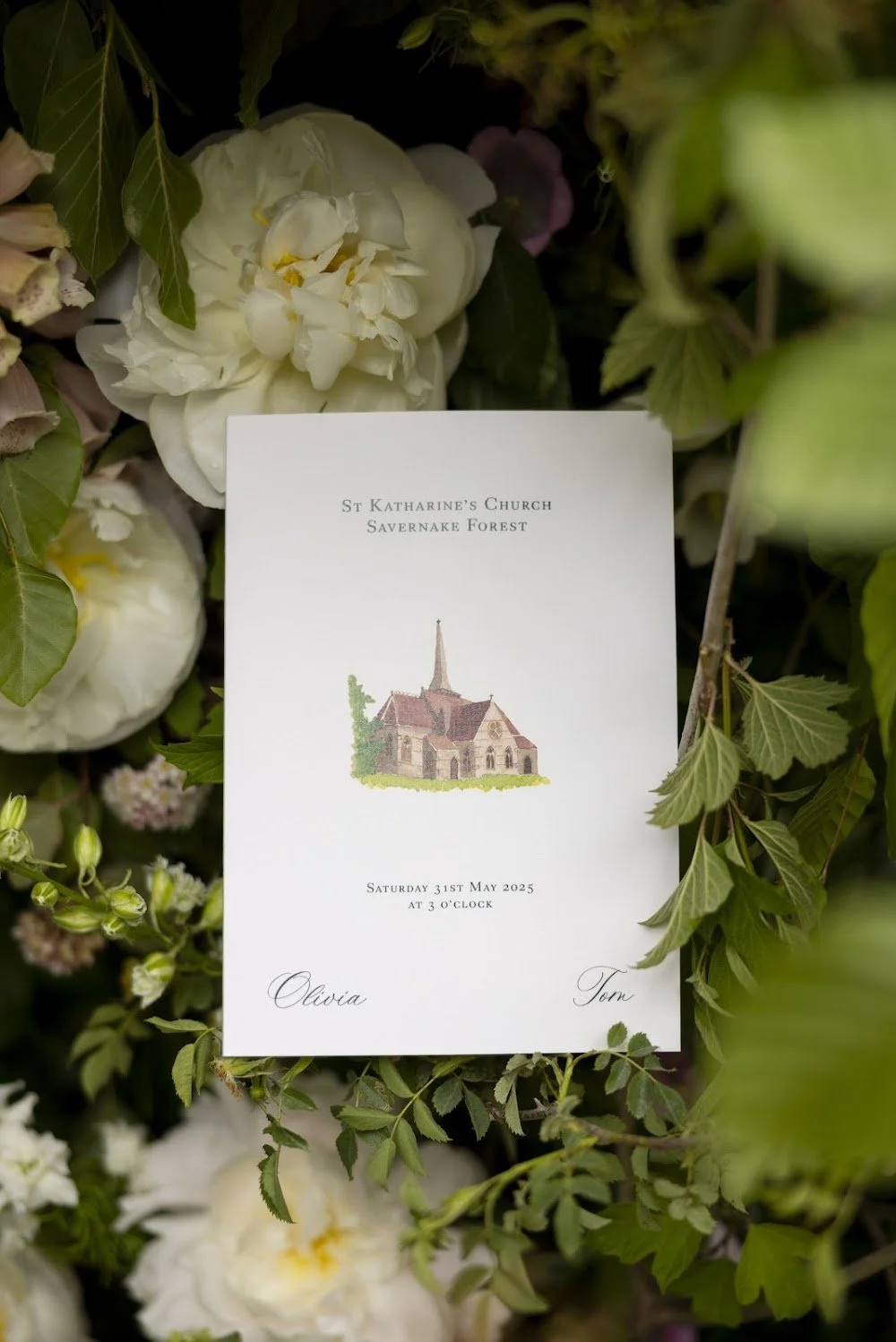 Wedding invitation card for St. Katharine's Church, Savernake Forest, surrounded by white and purple flowers and green foliage, with details of the date, time, and the names Olivia and Tom.