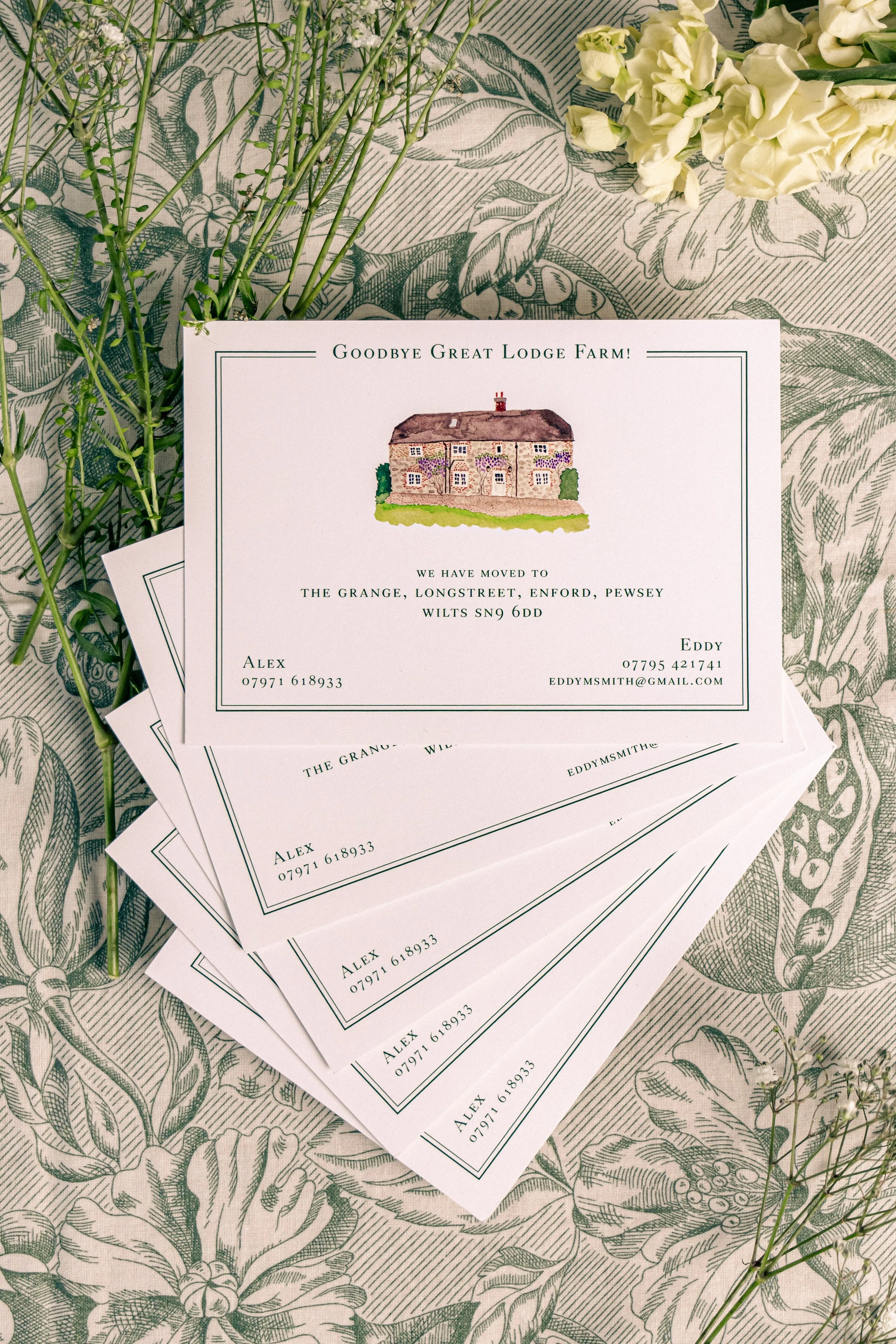 Stack of farewell cards with an illustration of a house, on a floral tablecloth with small white flowers nearby.