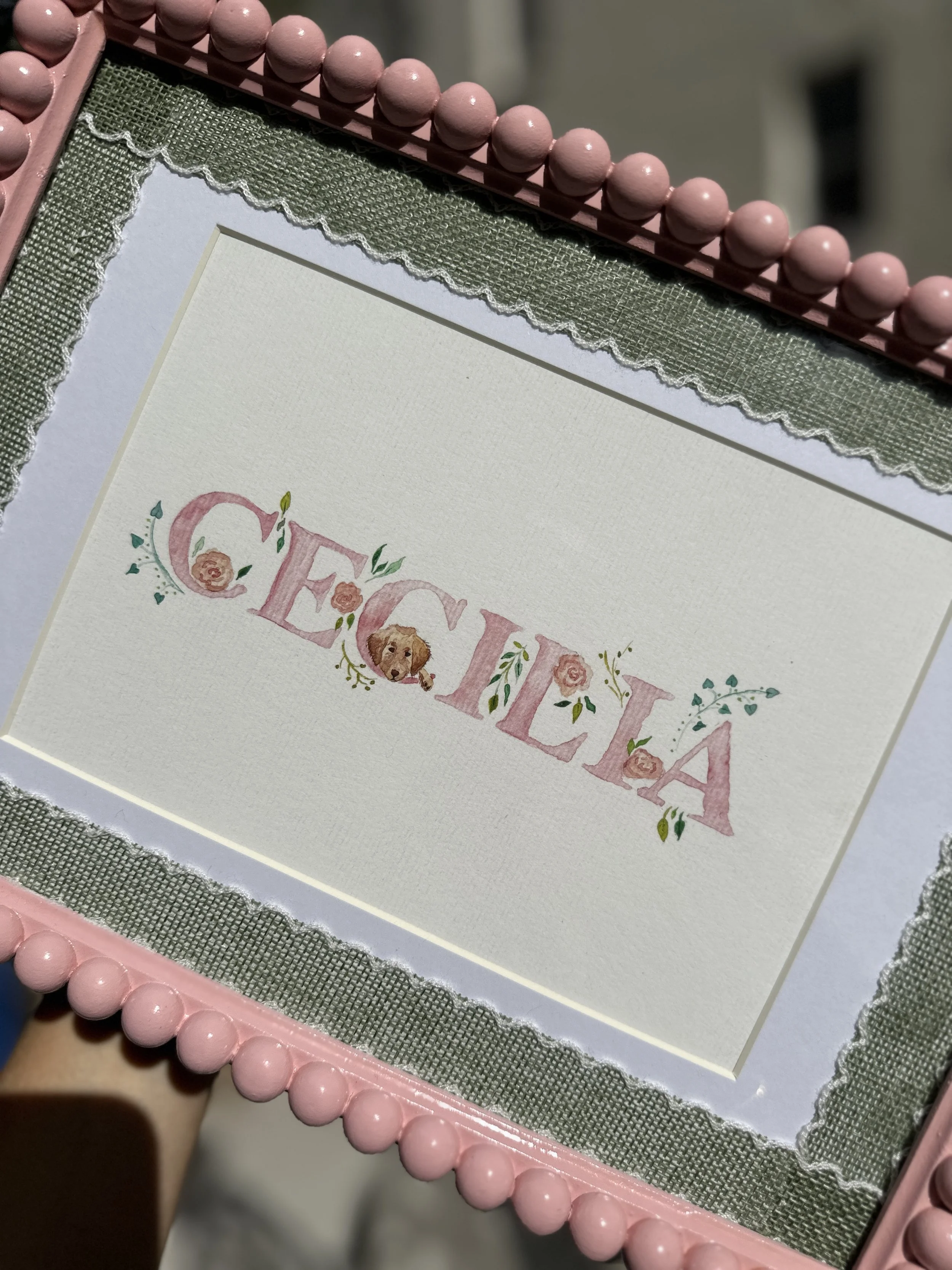 A decorative sign with the word 'FELICIA' written in pink, floral letters, featuring a dog illustration and surrounded by pink roses and green leaves.