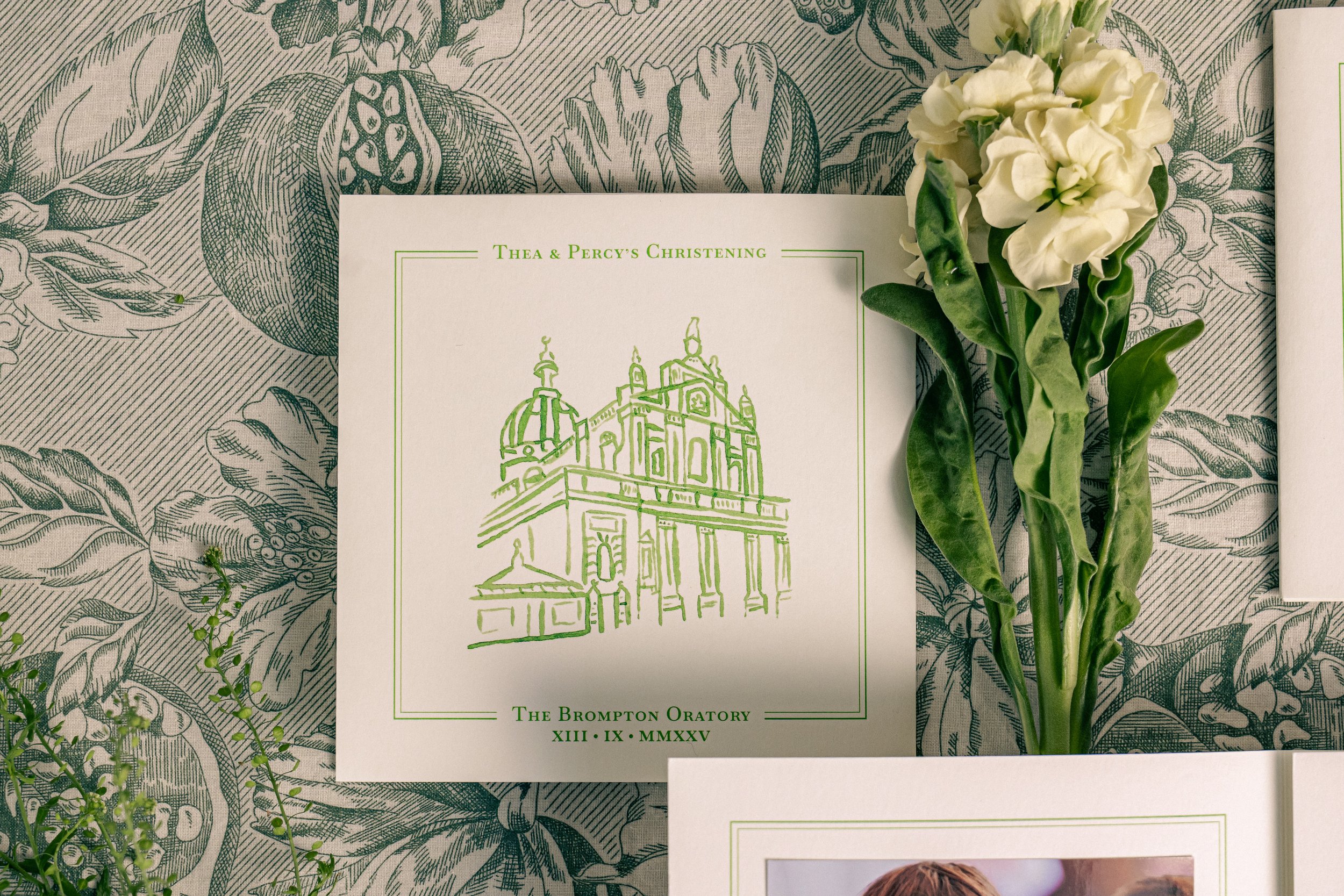 A wedding invitation card with green line art of the Brompton Oratory church, surrounded by white flowers and green leaves, on a botanical-patterned tablecloth.