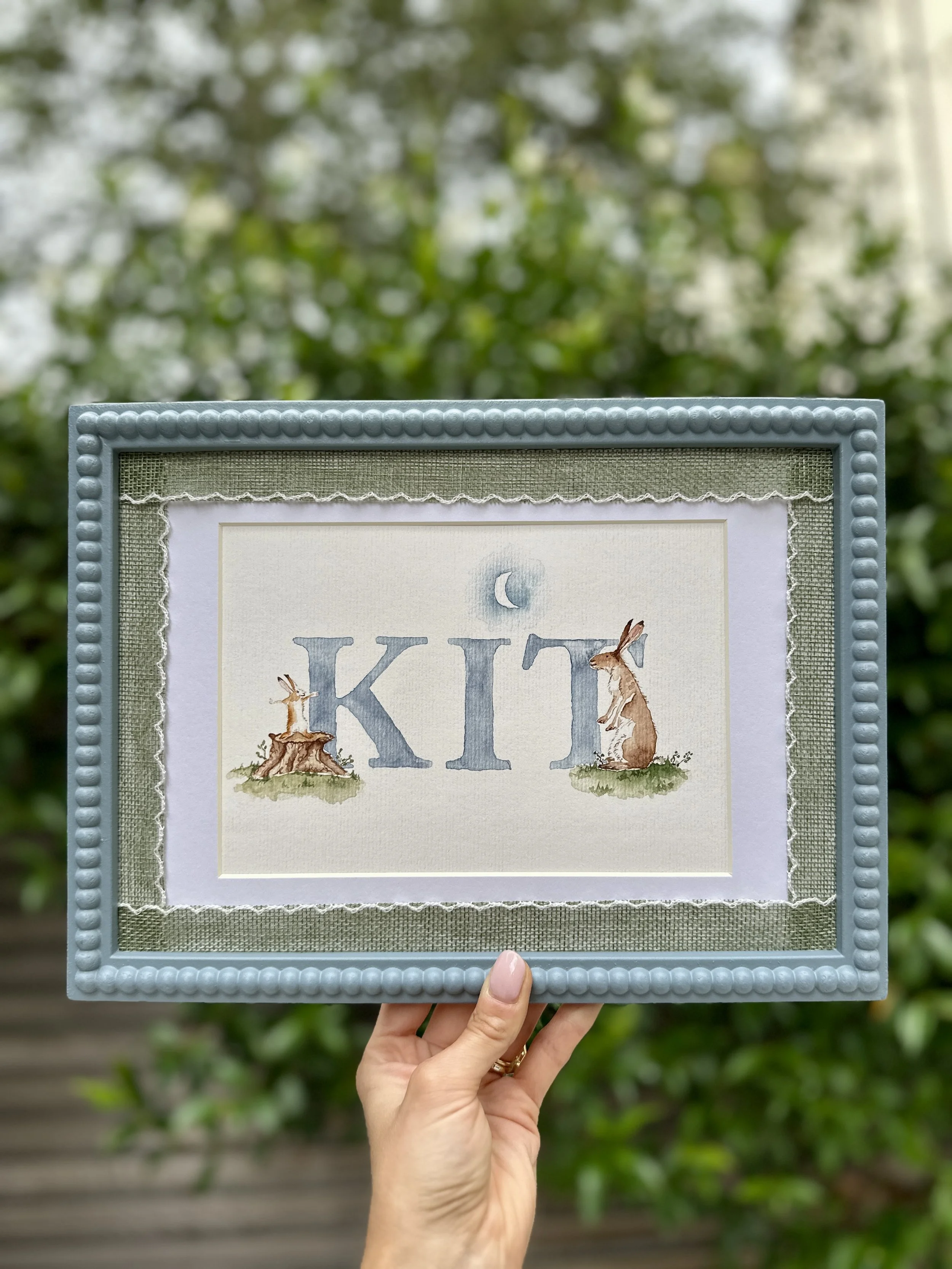 Decorative framed art with the word 'KID' in large blue letters, featuring illustrations of a rabbit and a hare, under a crescent moon, with a green textured background and a beaded border.