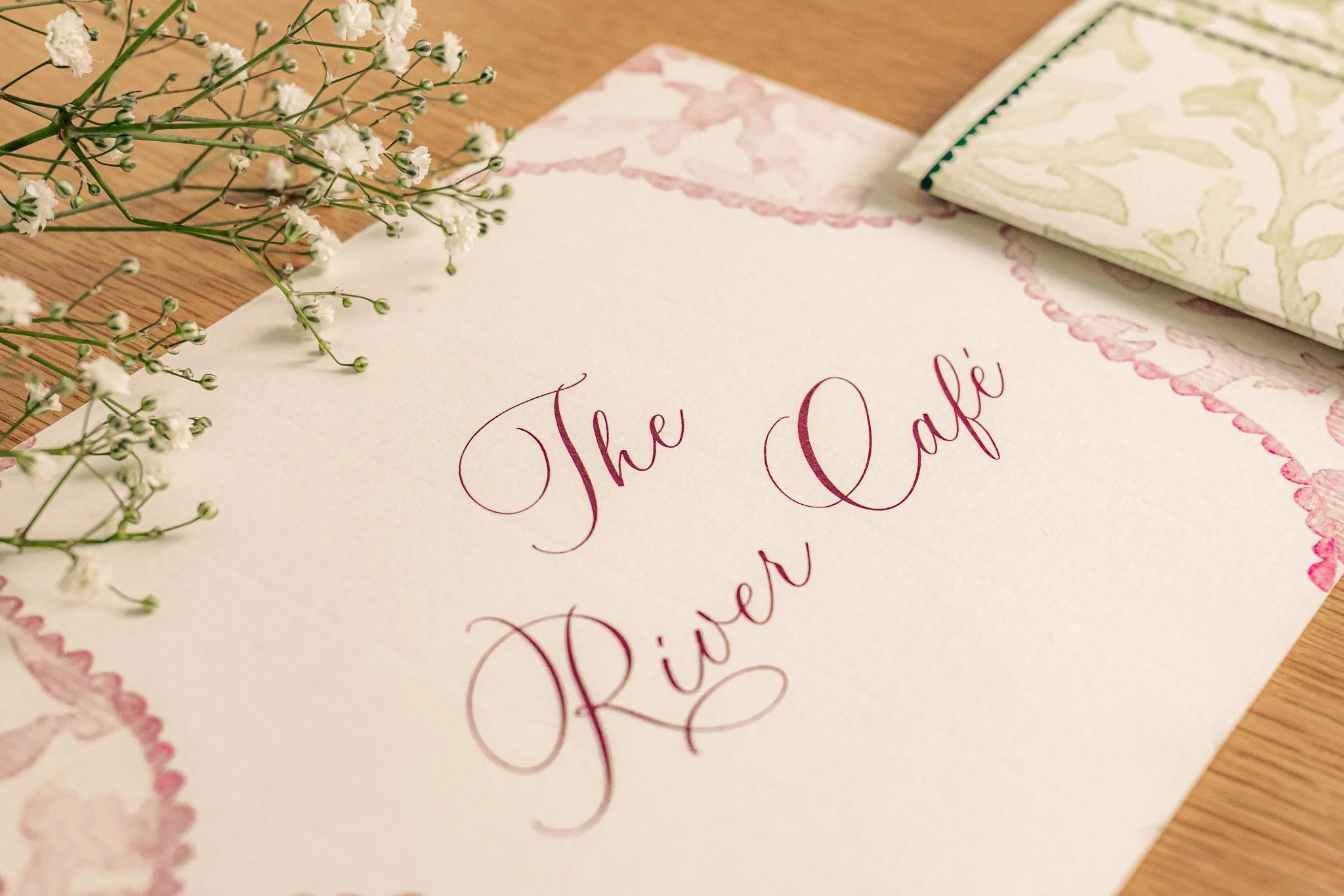 Card with pink cursive text reading 'The River Café' on a table with white flowers and patterned napkins.