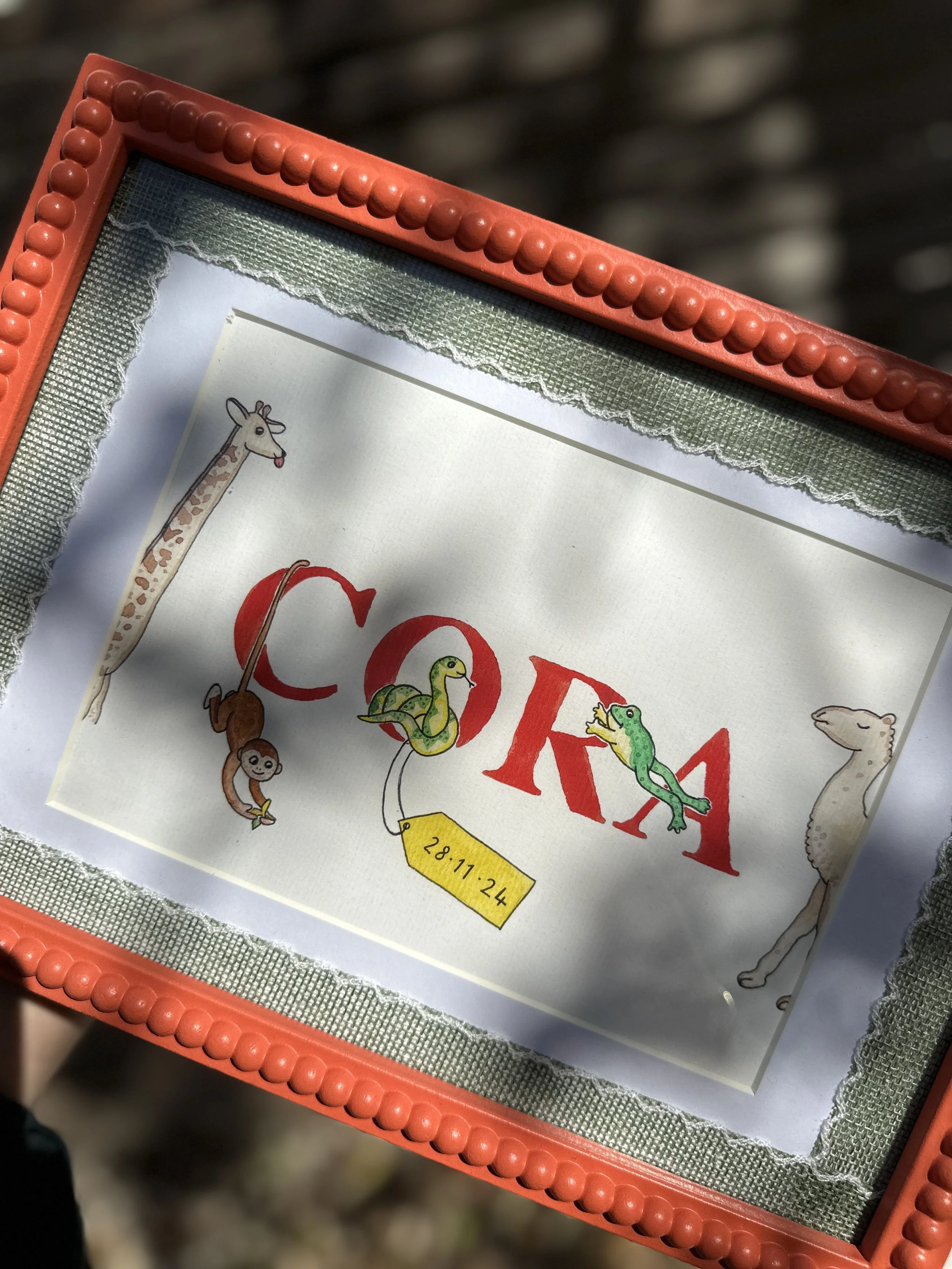 A framed artwork featuring the name 'Cora' with illustrations of a giraffe, monkey, snake, crocodile, and camel around the letters, and a yellow tag with the date 28.11.24.