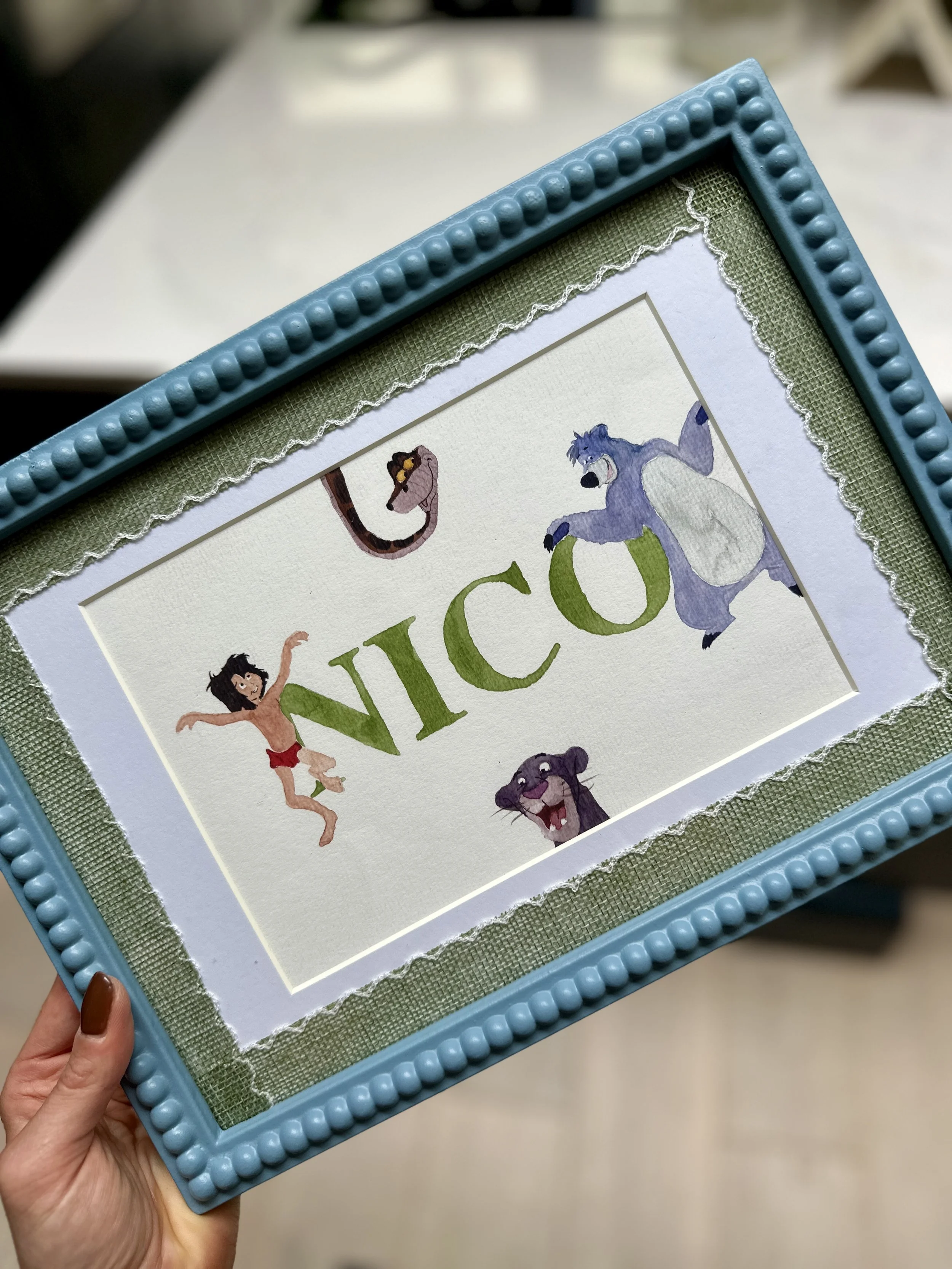 Hue watercolor painting of the character Mowgli from The Jungle Book, the character Baloo the bear, the snake Kaa, and the black panther Bagheera surrounding the name 'NICO' in green letters, all in a decorative blue frame.
