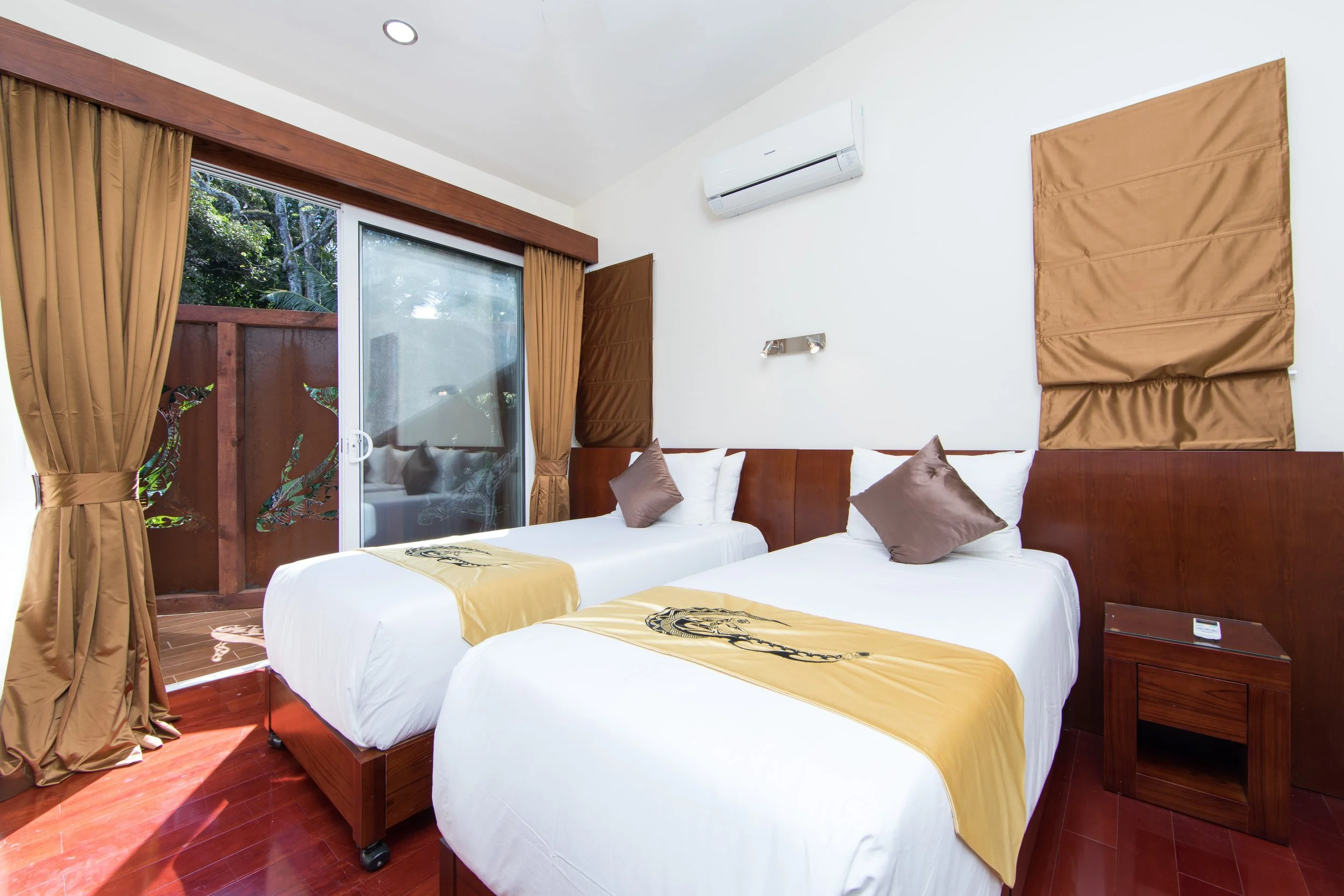 2 Bedroom Premium Villa second bedroom with two twin beds, white linens, brown pillows, wooden headboards, a sliding glass door with brown curtains, an ensuite, and small wooden nightstands.