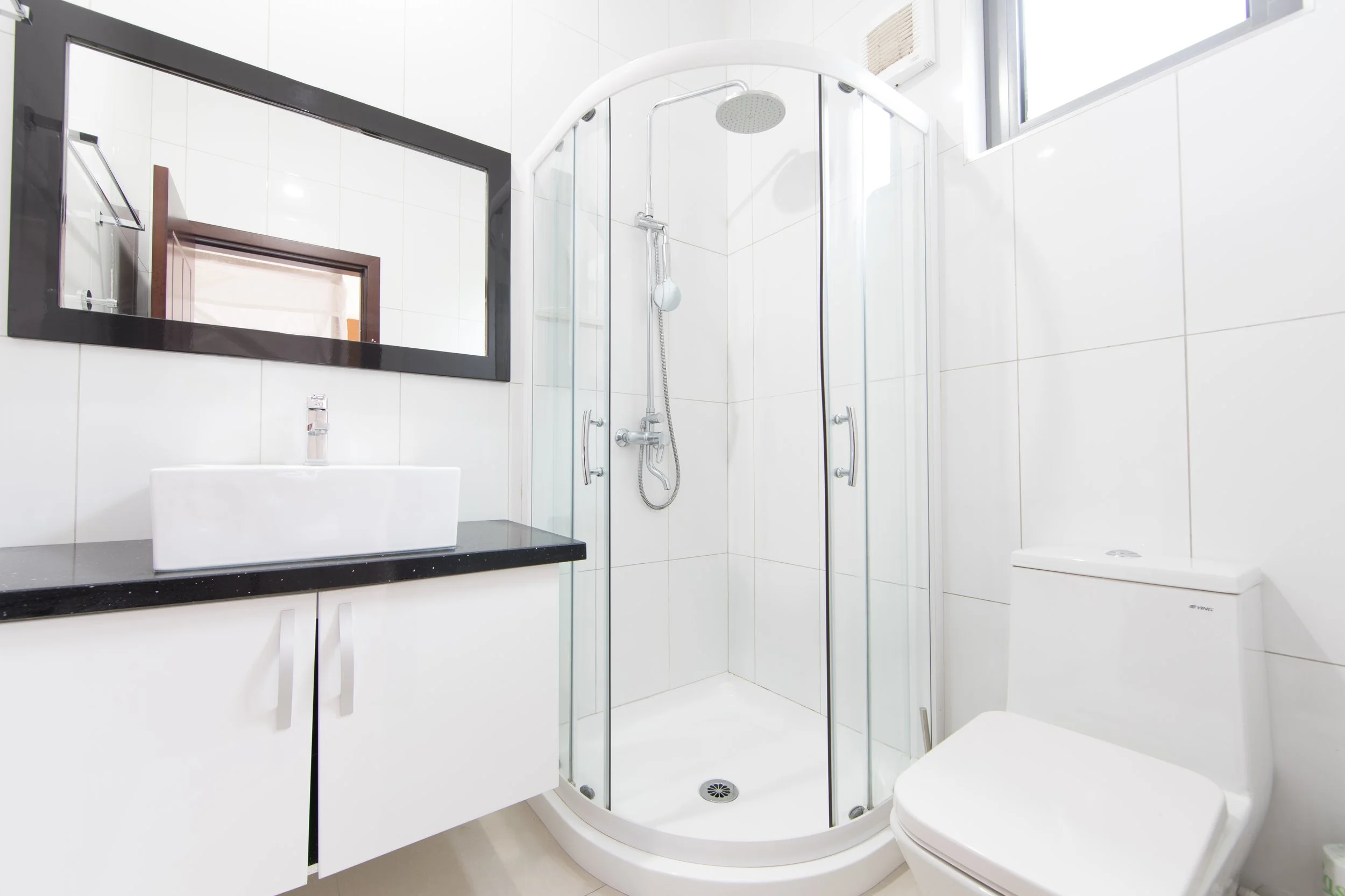 Honeymoon Villa bathroom with white tiled walls, a curved glass shower enclosure with a chrome showerhead, a white toilet, a black countertop with a white rectangular sink, and a large rectangular mirror.