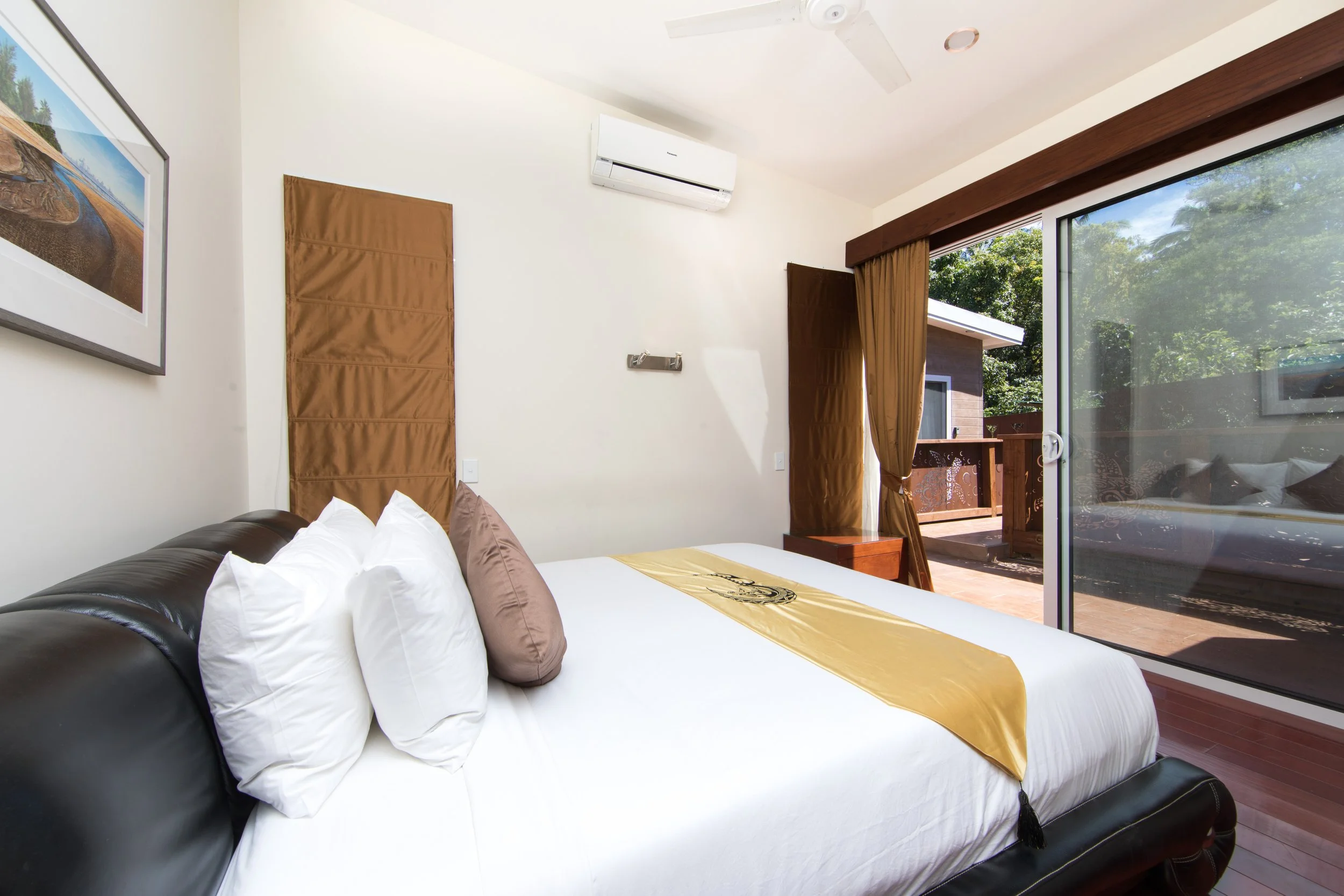 2 Bedroom Premium Villa main bedroom with a king leather bed, bedding, and pillows, near a sliding glass door leading to an outdoor balcony with tree views outside.
