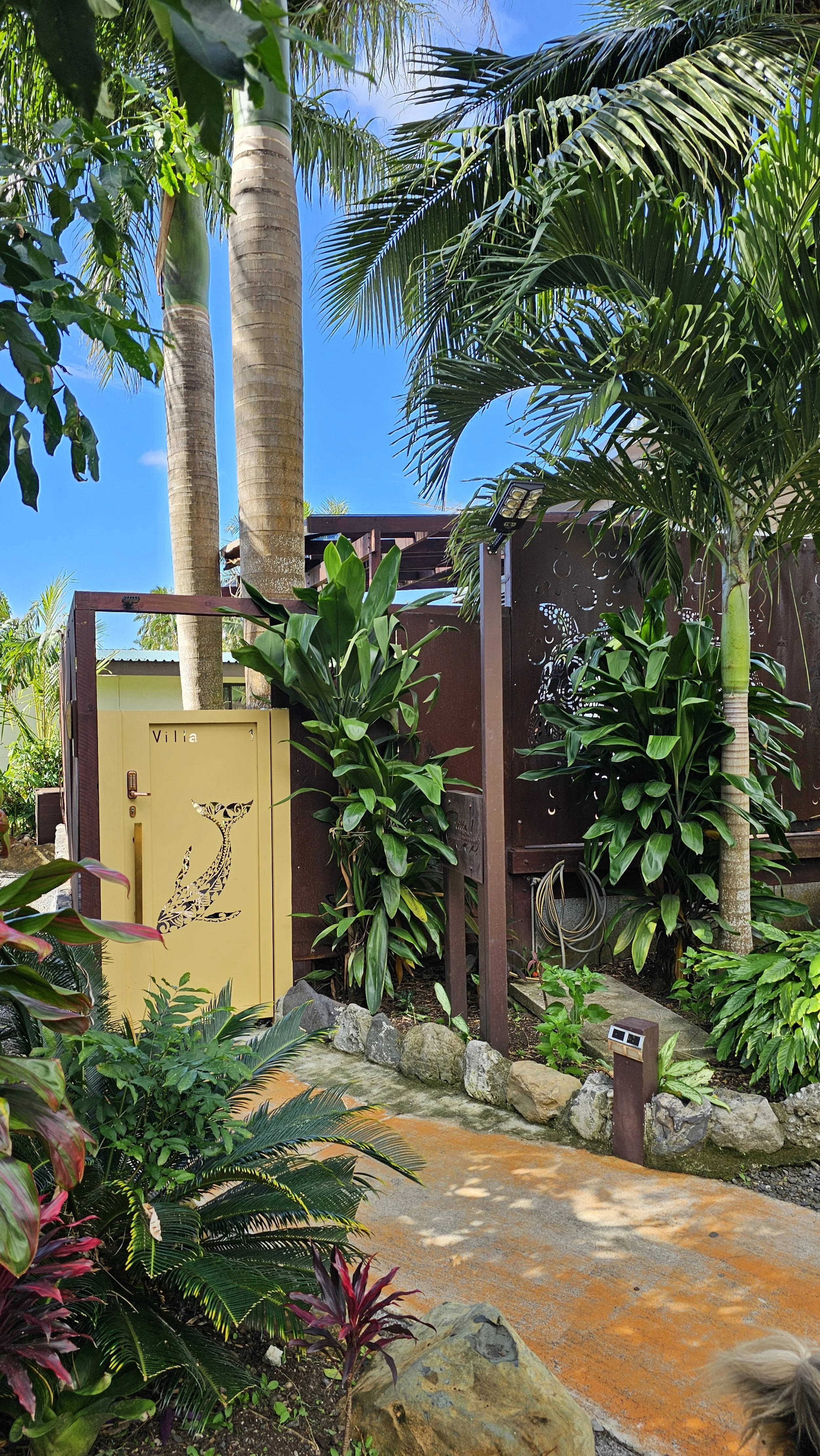 Honeymoon Villa entrance. A tropical garden scene with tall palm trees, lush green plants, a yellow door with the word "Villa" and a decorative bird silhouette, a stone border, a pathway, and a blue sky in the background.