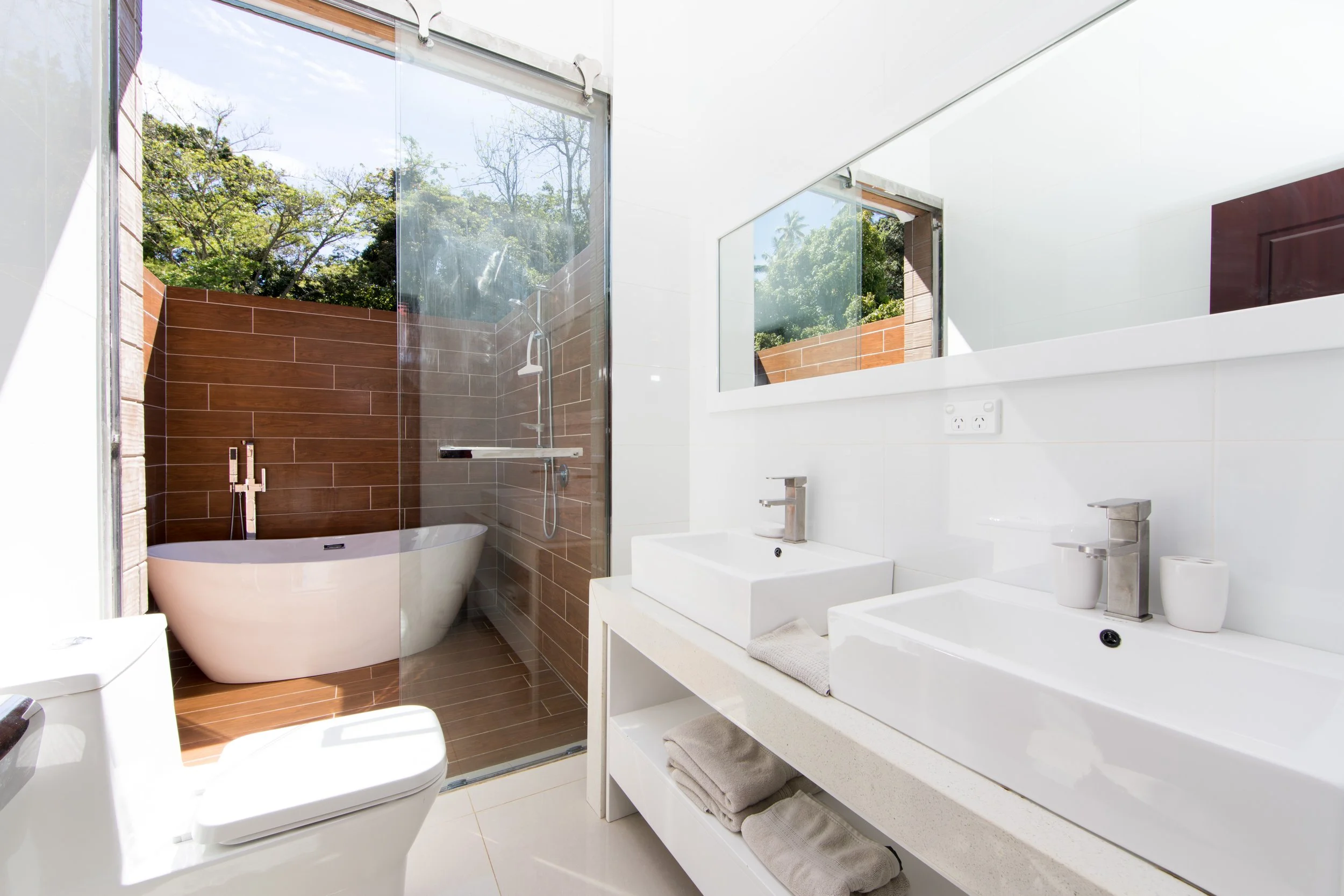 2 Bedroom Premium Villa modern ensuite with double sinks, a large mirror, white fixtures, and a glass-enclosed shower with a freestanding tub outside, with views of greenery.