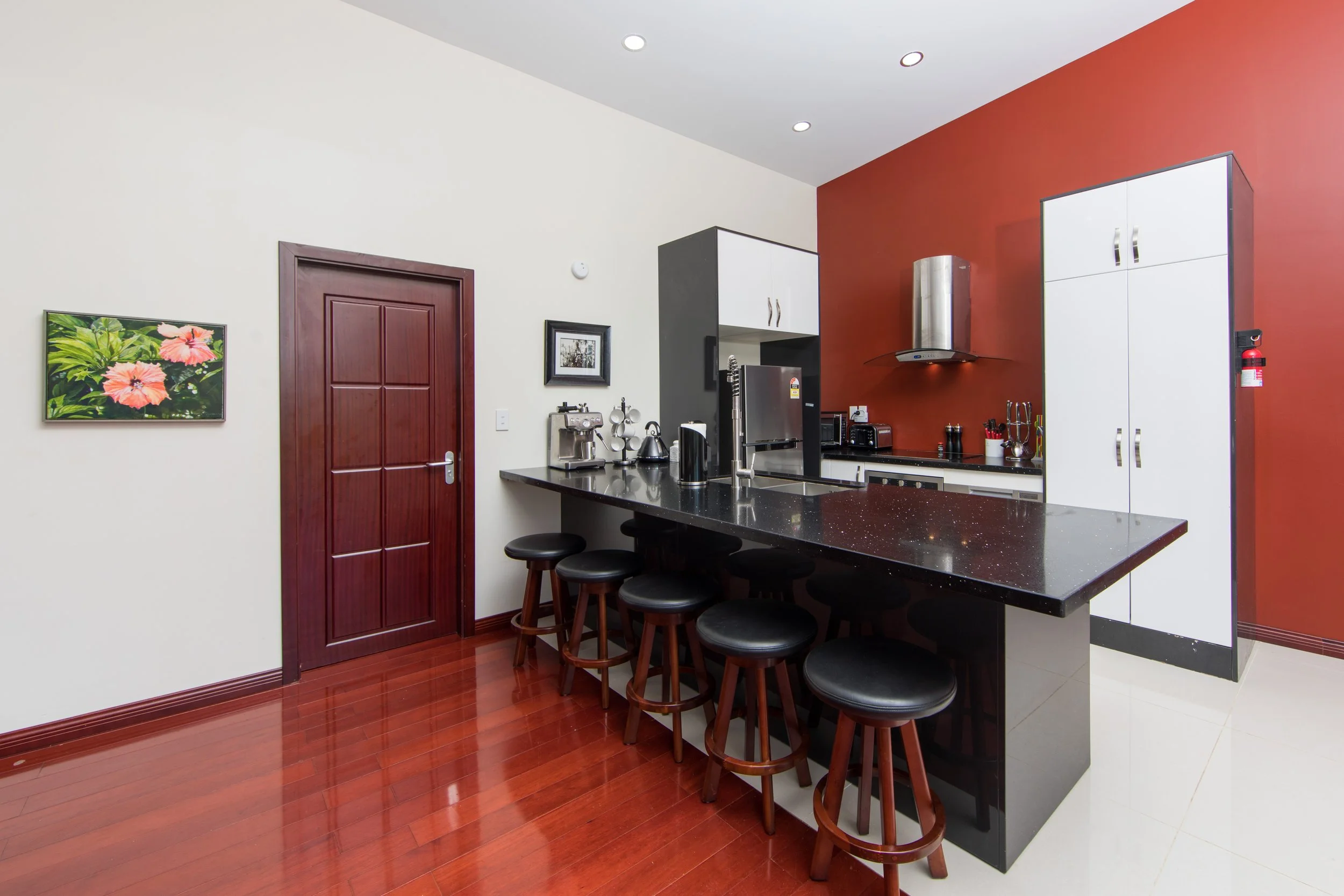 2 Bedroom Premium Villa - Modern kitchen with white and dark gray cabinetry, a red accent wall, black granite countertop with bar stools, stainless steel appliances, and hardwood and tile flooring.