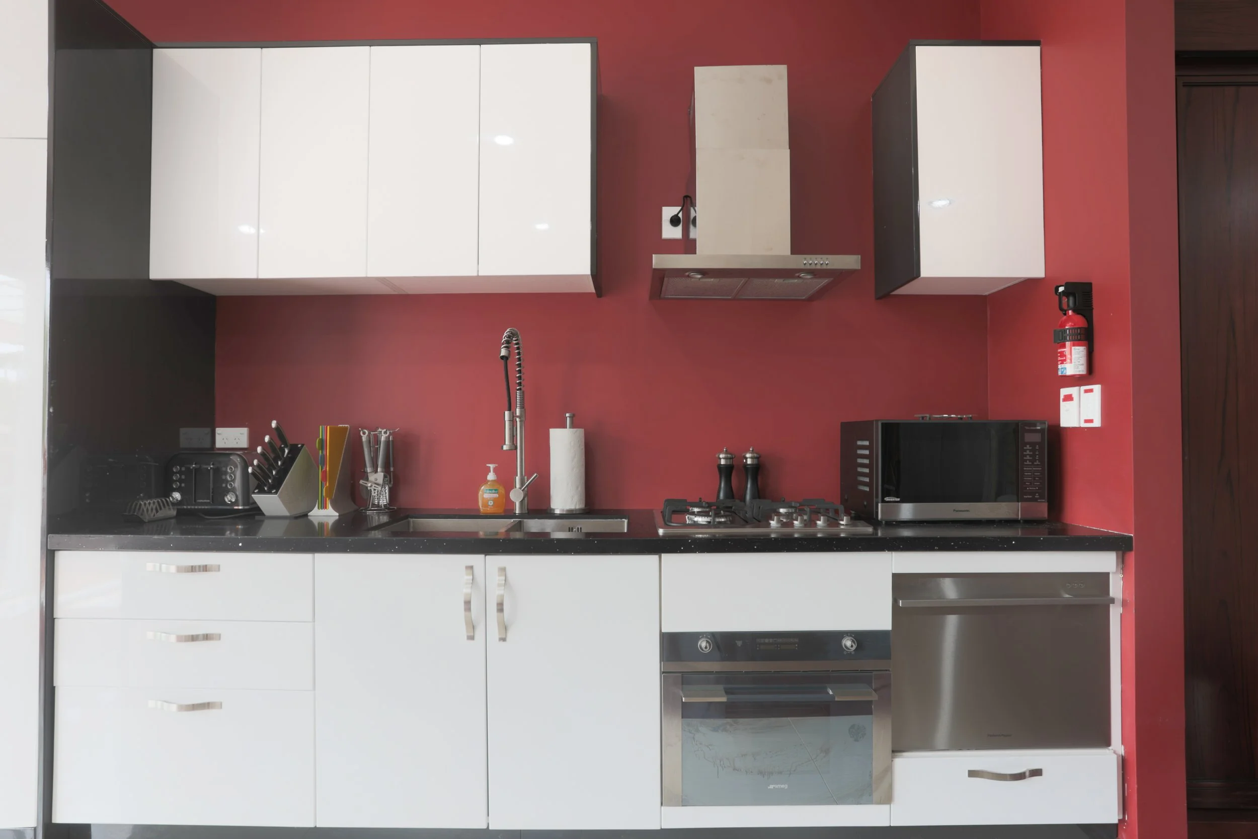 Garden Villa modern kitchen with red accent wall, white upper and lower cabinets, black granite countertop, stainless steel appliances including oven, microwave, and range hood, and various small appliances on the counter.