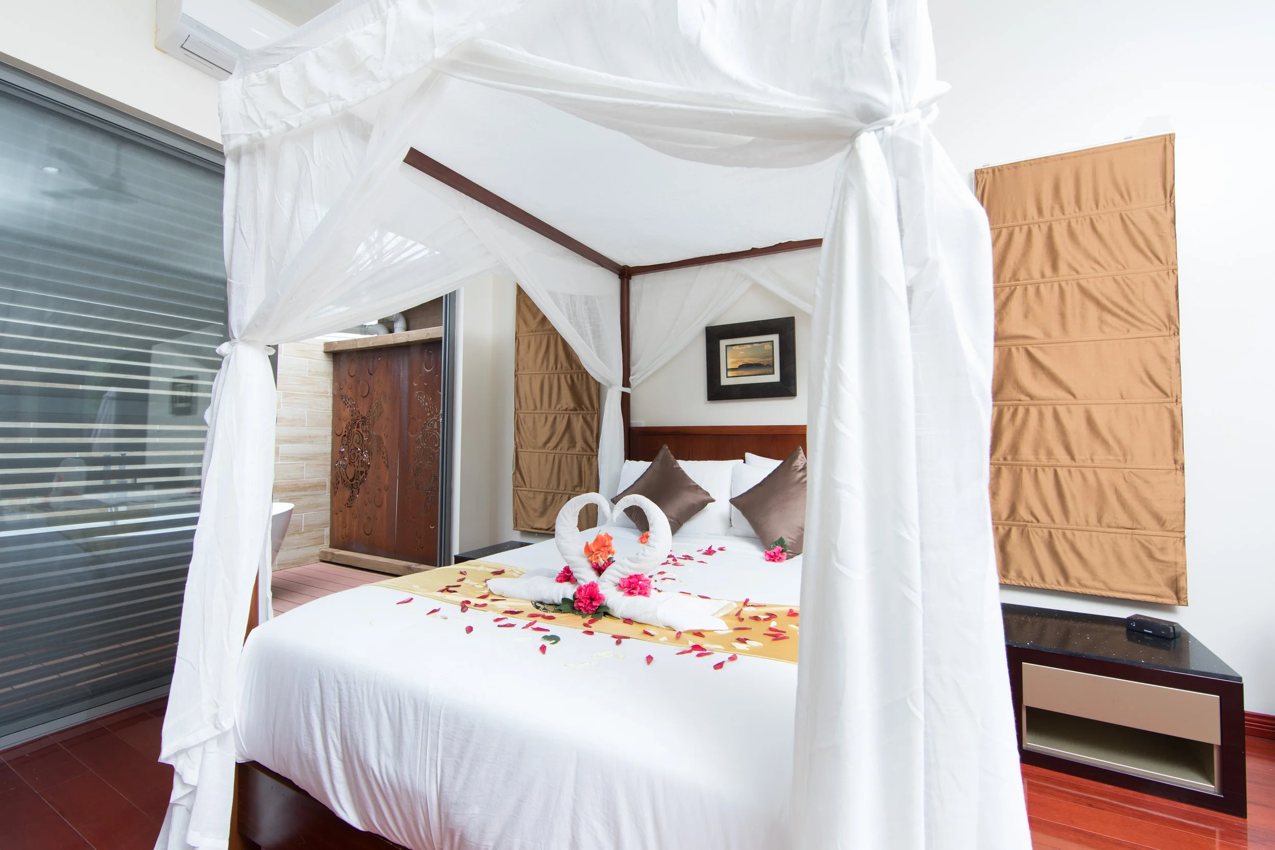 Honeymoon Villa four-poster bed decorated with swan-shaped towels, flower petals, and pillows, with a small nightstand and artwork on the wall.