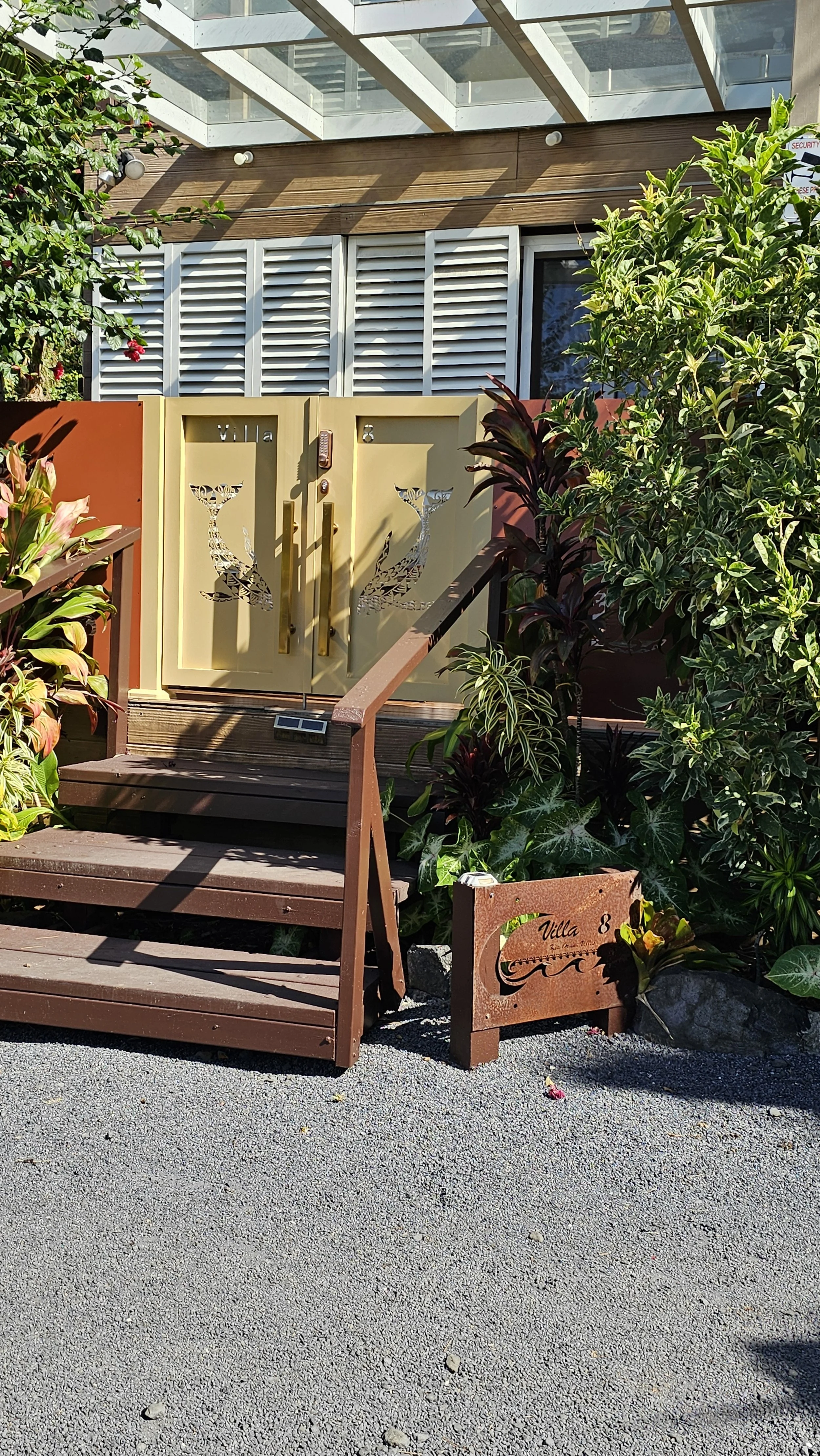 Garden Villa entrance with a yellow gate decorated with whale cutouts, surrounded by green plants and bushes, with steps leading up to a building with white shutters and glass roof.