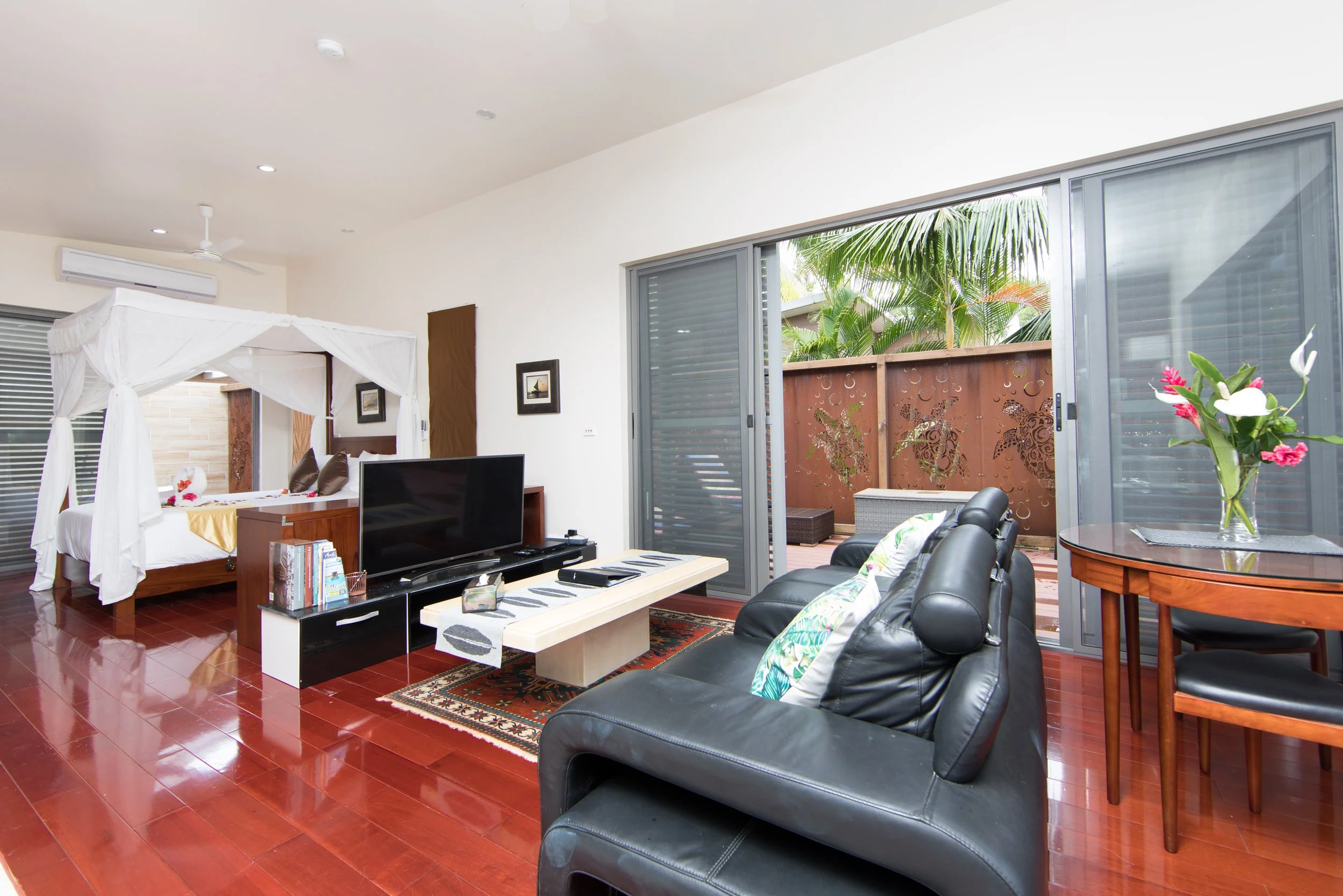 Honeymoon Villa lounge with a four-poster bed, a flat-screen TV, a leather sofa, and a sliding glass door leading to a balcony with tropical plants.