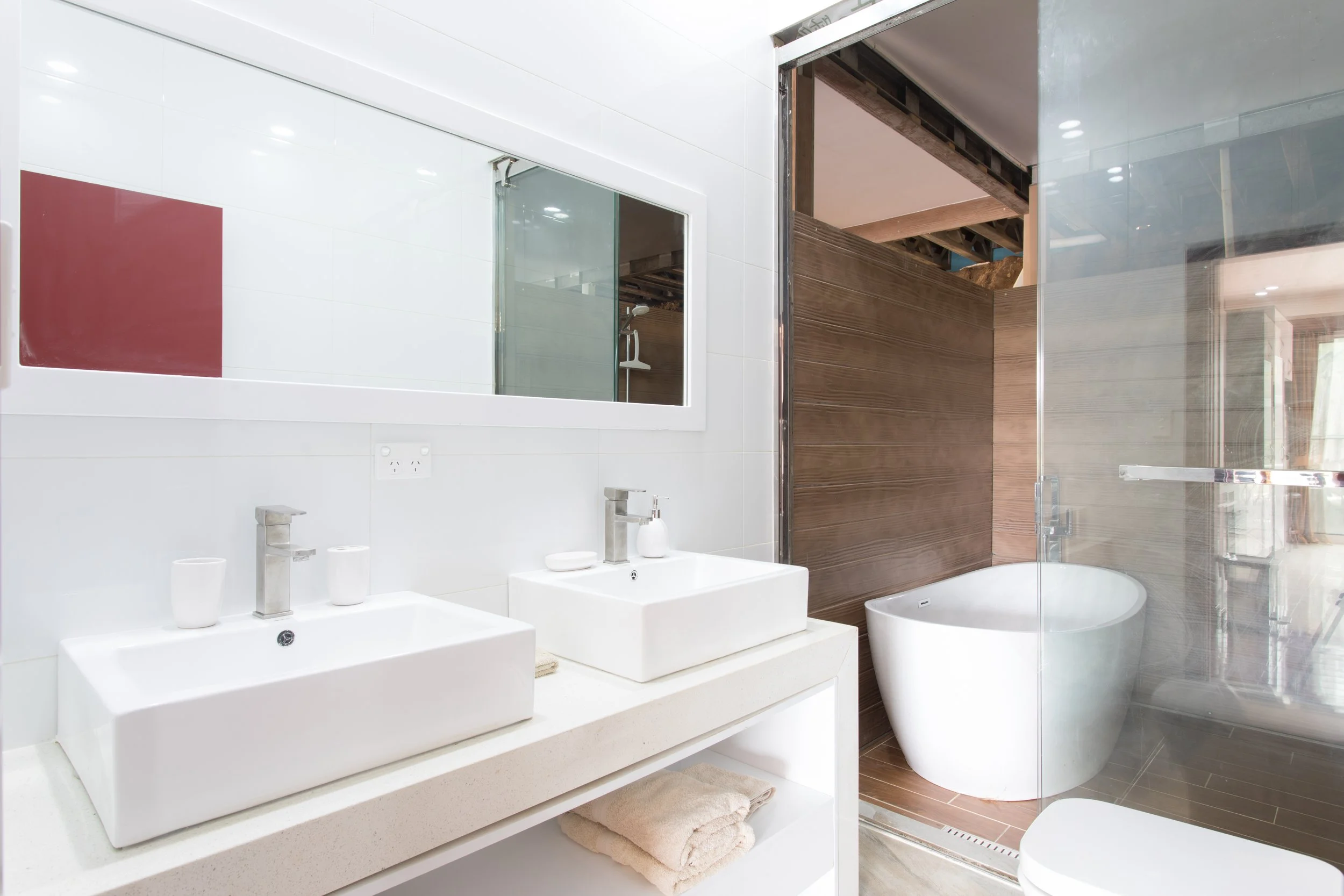 Garden Villa modern bathroom with white double sinks, a large mirror, a towel, and a separate glass-enclosed bathtub area with wooden walls.