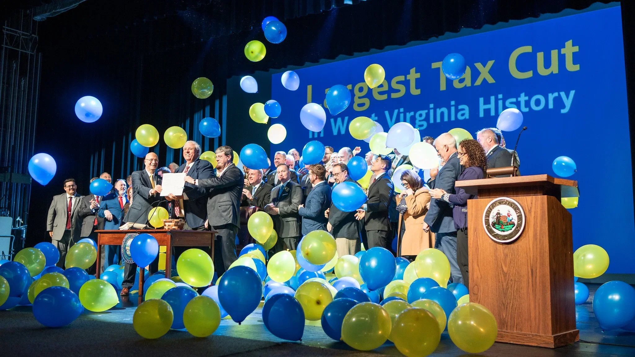 West Virginia state officials celebrating at a ceremony with balloons and a large screen displaying 'Largest Tax Cut in West Virginia History.'