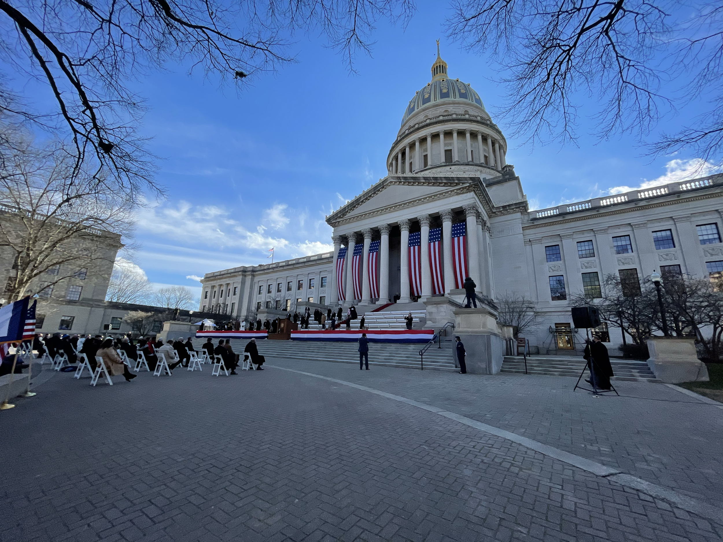 The West Virginia Capitol building decorated with American flags and red, white, and blue banners, with an outdoor event setup on the steps and attendees seated in chairs facing the stage.