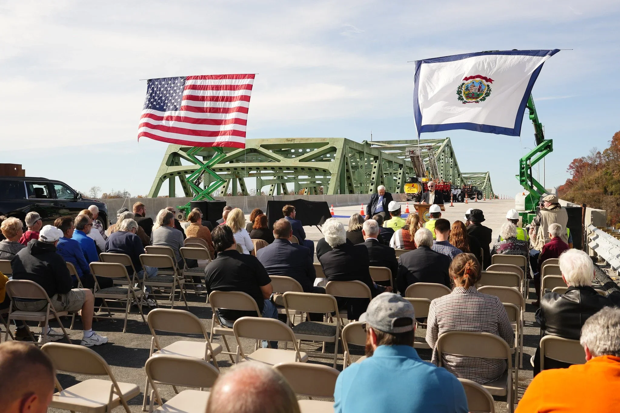 A crowd attending an outdoor event on a bridge with American and state flags, with speakers at a podium and construction equipment in the background.