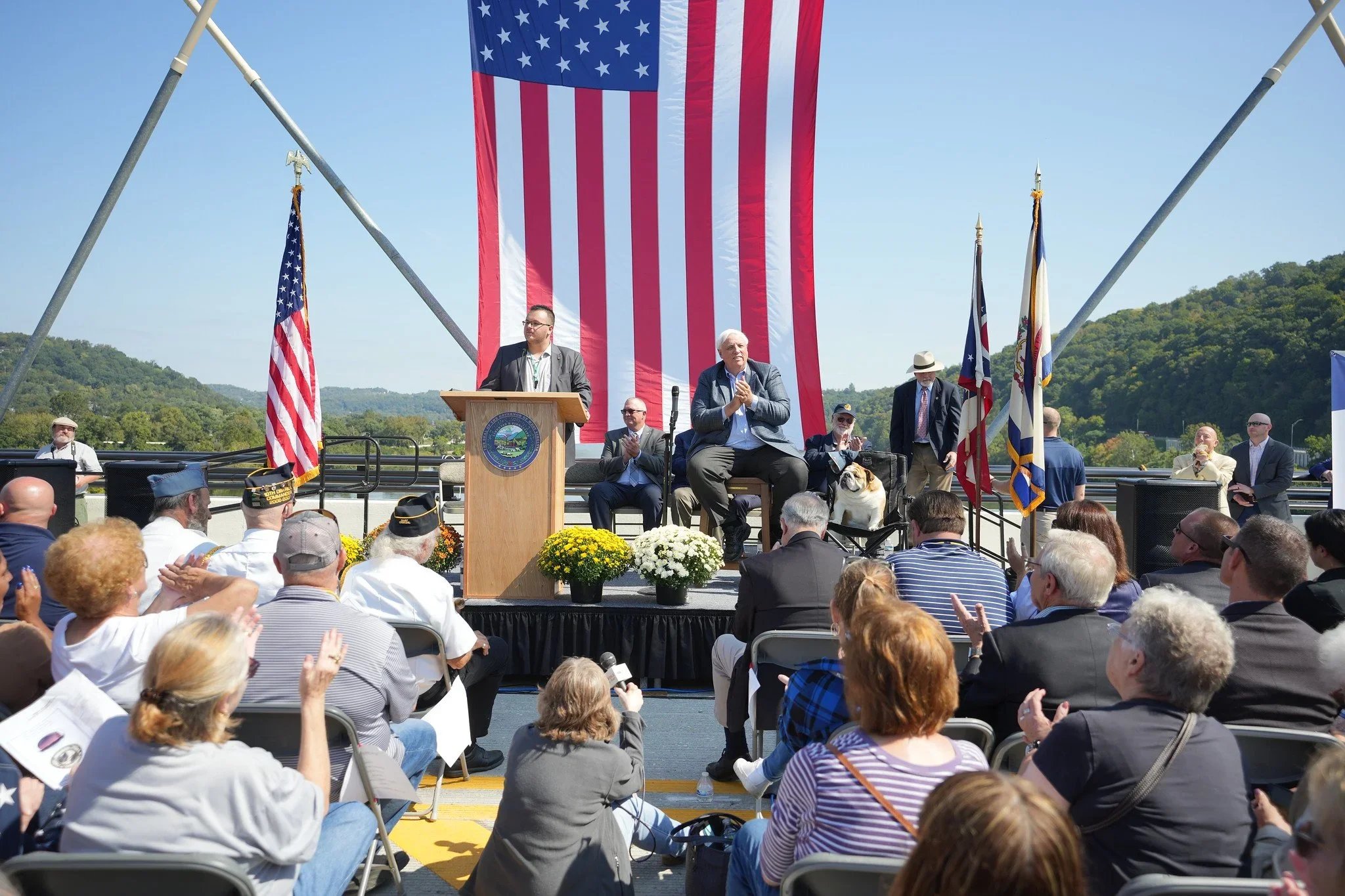 A patriotic outdoor event with a large American flag hanging above a stage. Several people are on stage, including a man at a podium, some sitting, and others standing with flags in the background. The audience is engaged, some clapping, with mountai