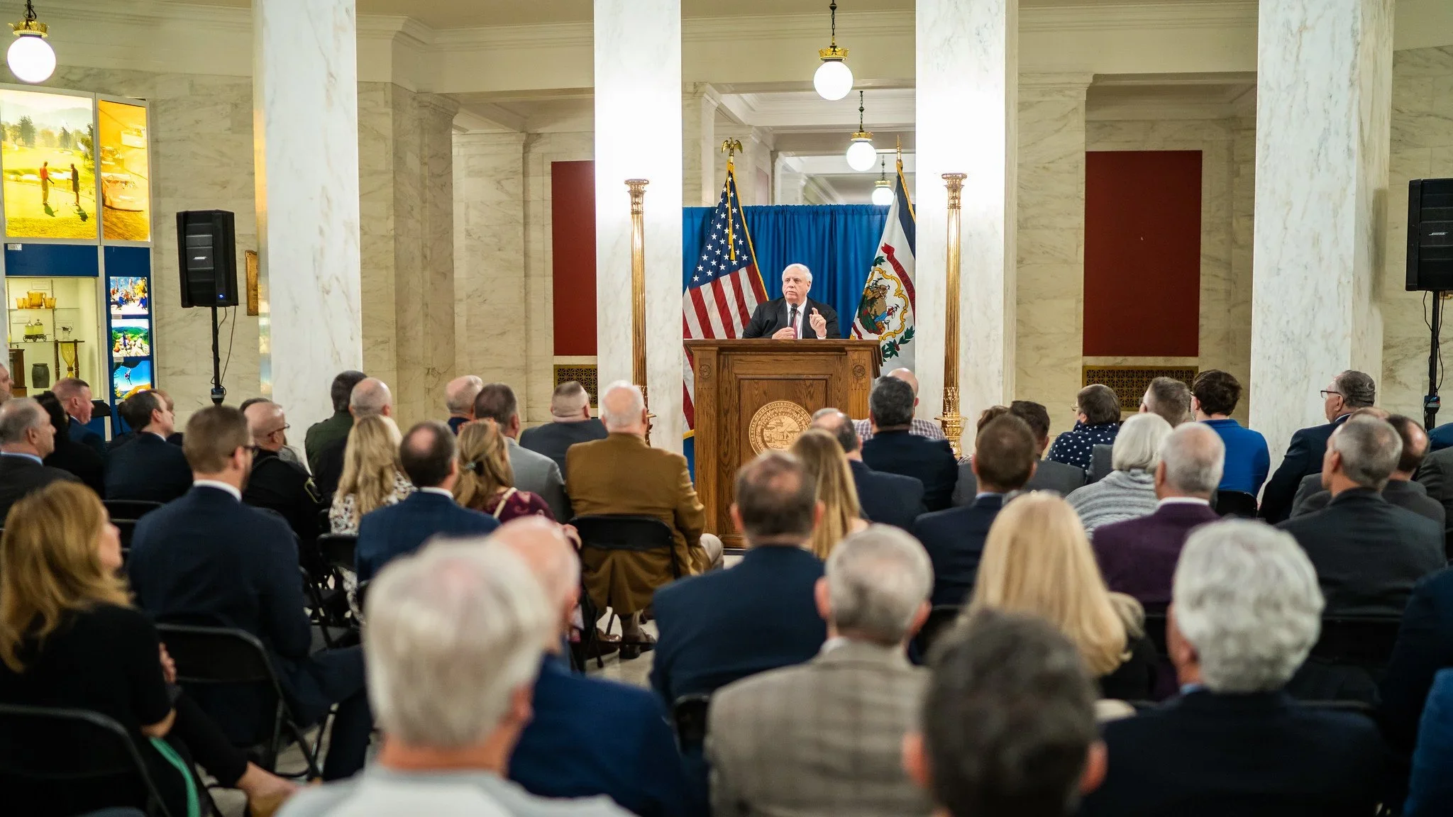 A man giving a speech at a podium during a formal event, with American and state flags behind him, in a large, marble-floored hall with an audience seated facing him.
