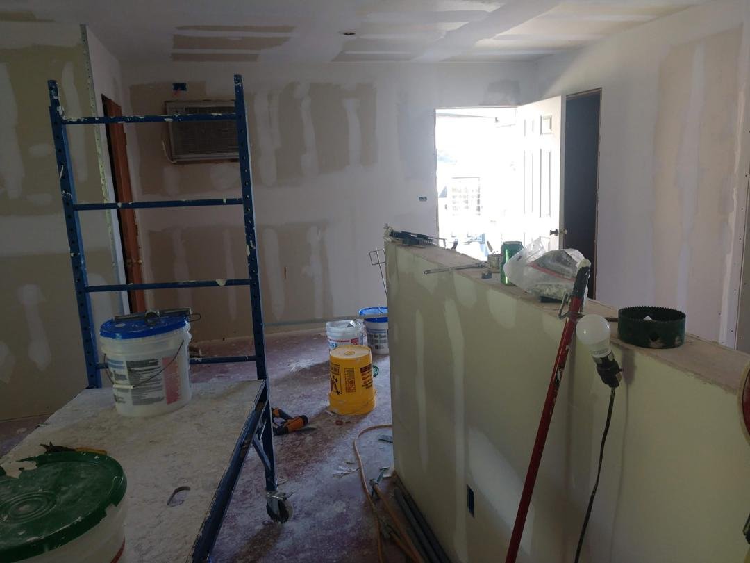 Room under renovation with partially painted drywall, construction tools, and materials including buckets, paint rollers, a ladder, and a partially built wall.