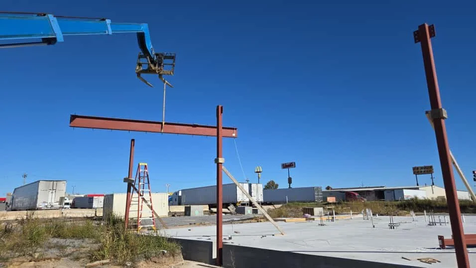 Steel beams assembled on a construction site with a crane arm overhead, and construction materials and trailers in the background on a clear day.