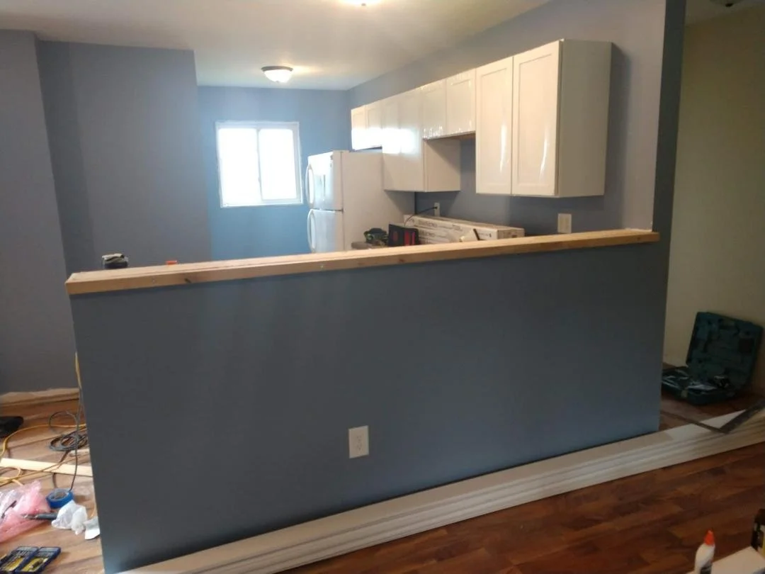 Partially renovated kitchen with blue walls, white cabinets, a window, a refrigerator, and a half-wall with a wooden top, construction tools, and materials on the floor.