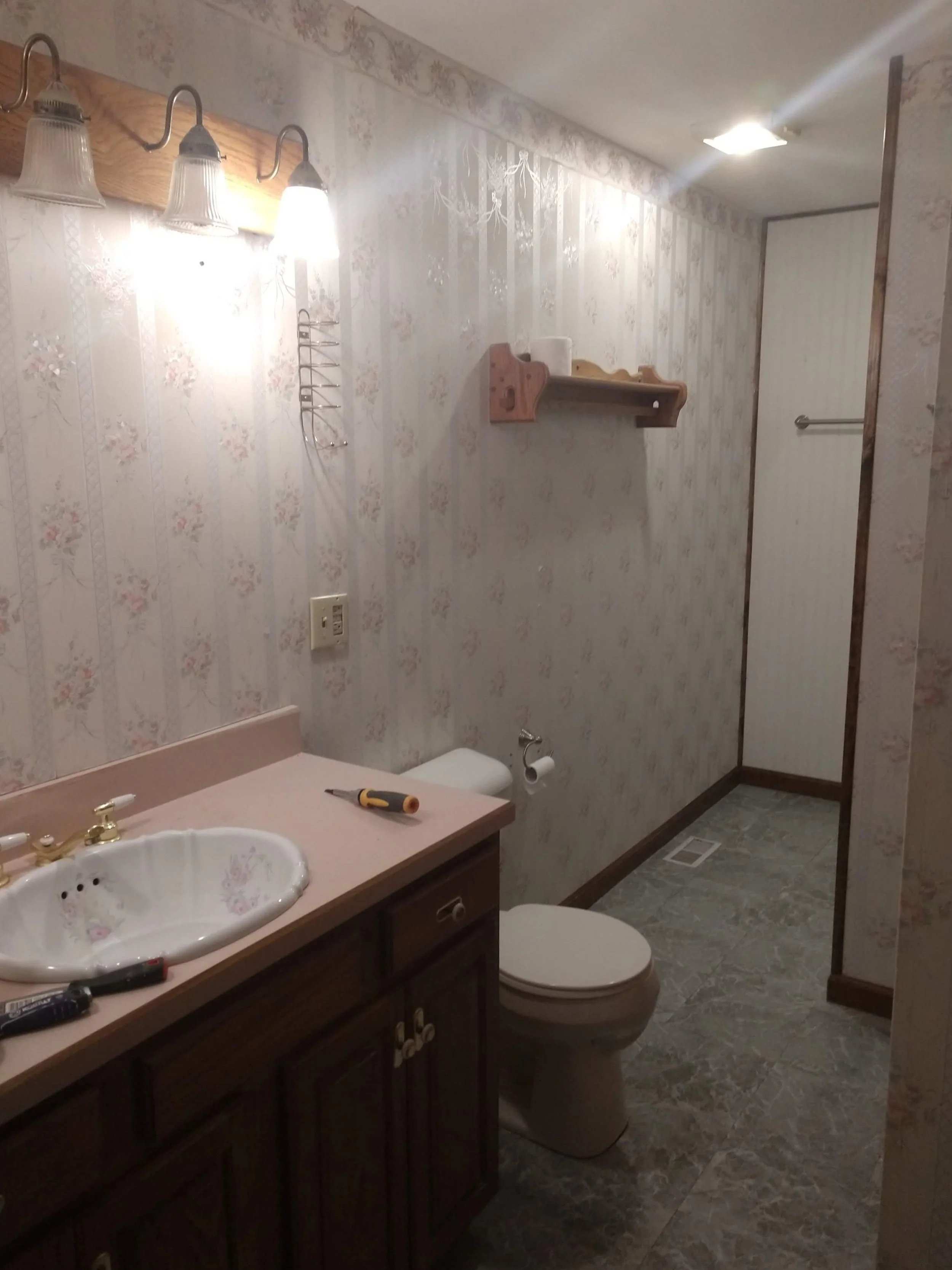 A small bathroom with floral wallpaper, a wooden cabinet with a pink countertop, a white sink with gold fixtures, a toilet with a toilet paper roll, a wall-mounted wooden shelf, and a light fixture with four glass shades. There are tools on the count