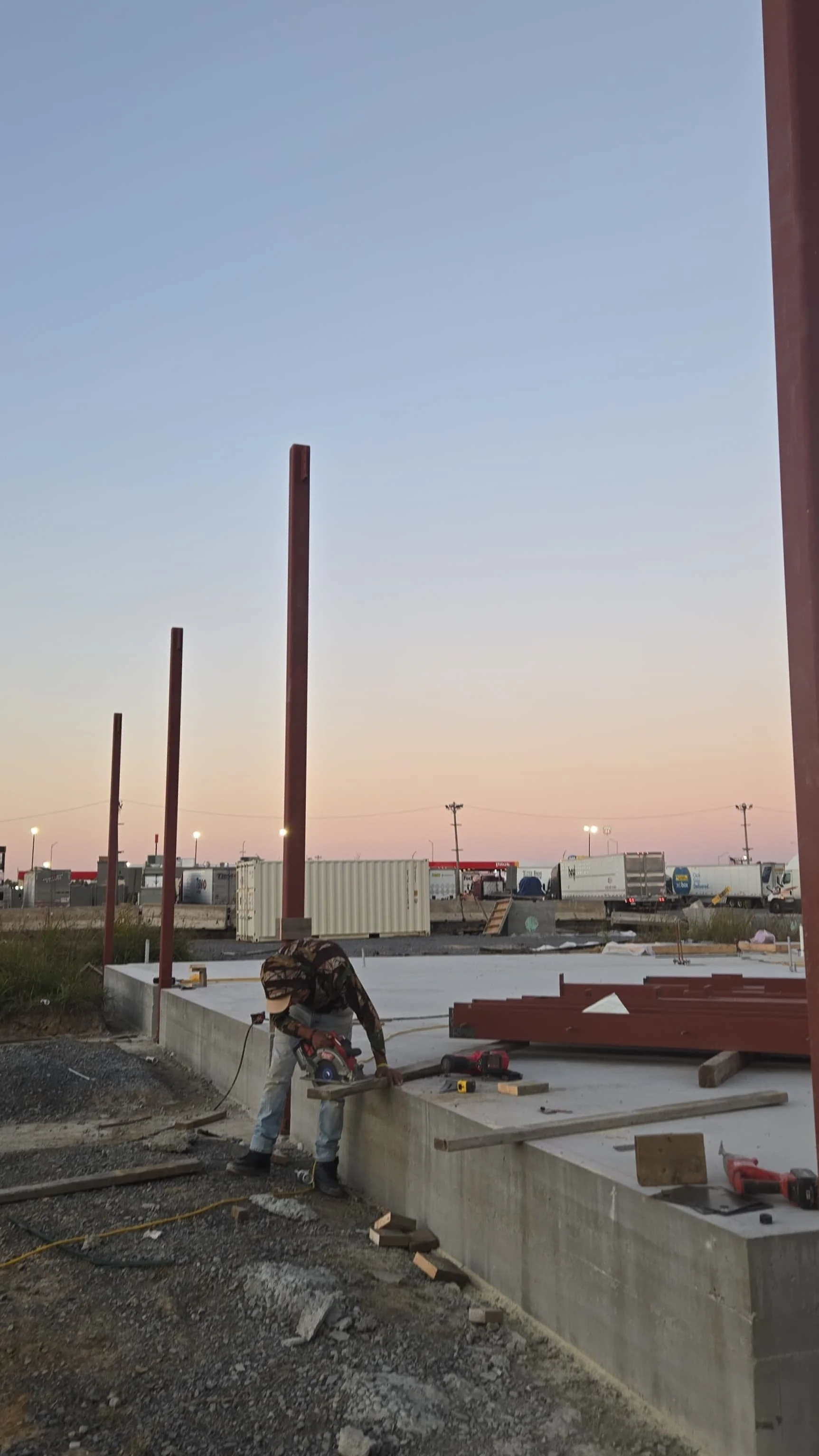 A construction worker cutting a piece of wood on a concrete foundation at a construction site during sunset, with vertical steel posts in the background and trucks parked nearby.
