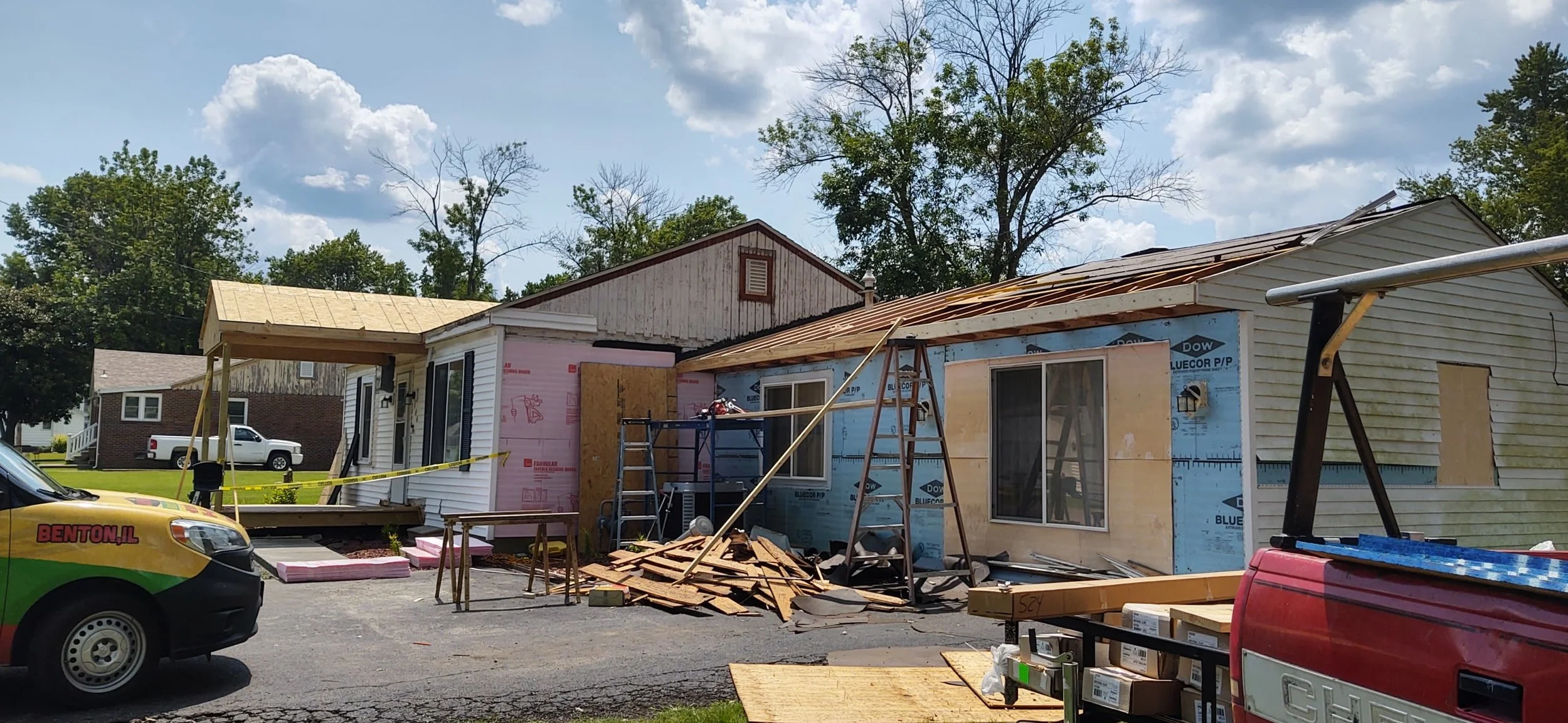 House under renovation with construction materials and tools around, including ladders, wood planks, and trucks, with partly replaced roof and ongoing siding work.