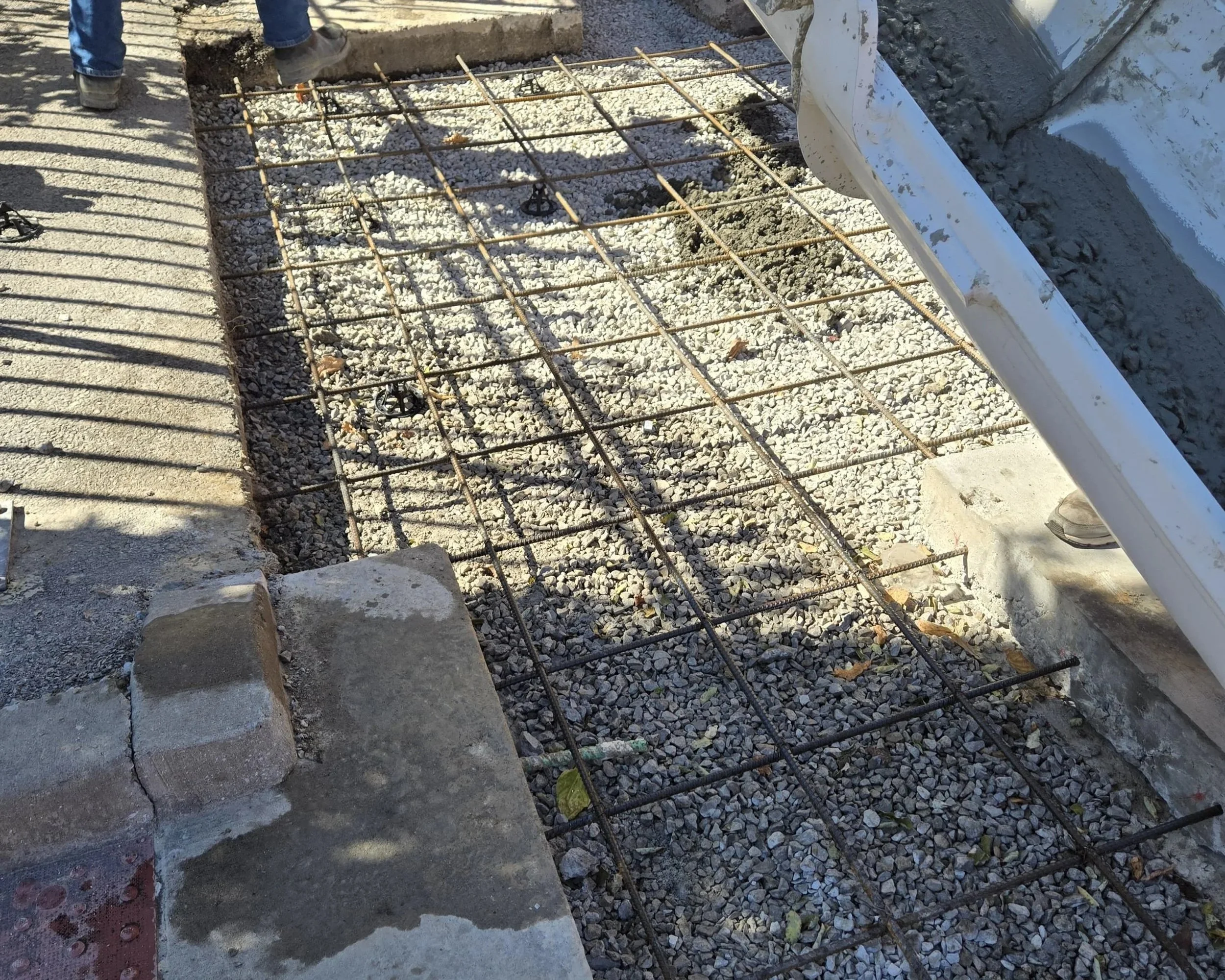 Construction site with steel rebar grid for concrete pouring, gravel ground, and a nearby portable cement mixer.