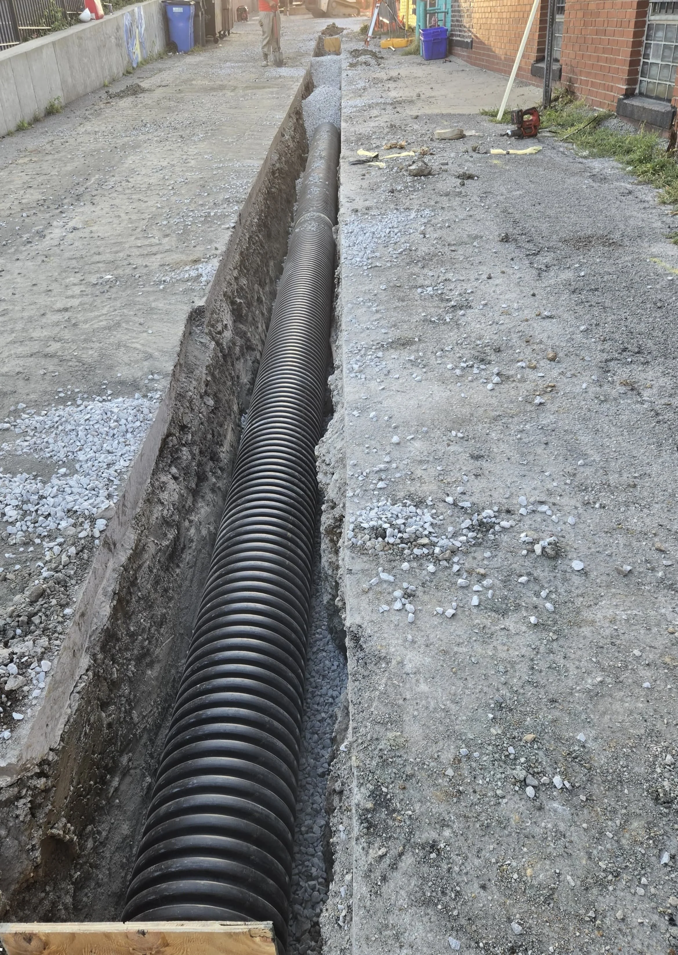 A construction site on a sidewalk where a large black corrugated pipe is being installed in a trench.