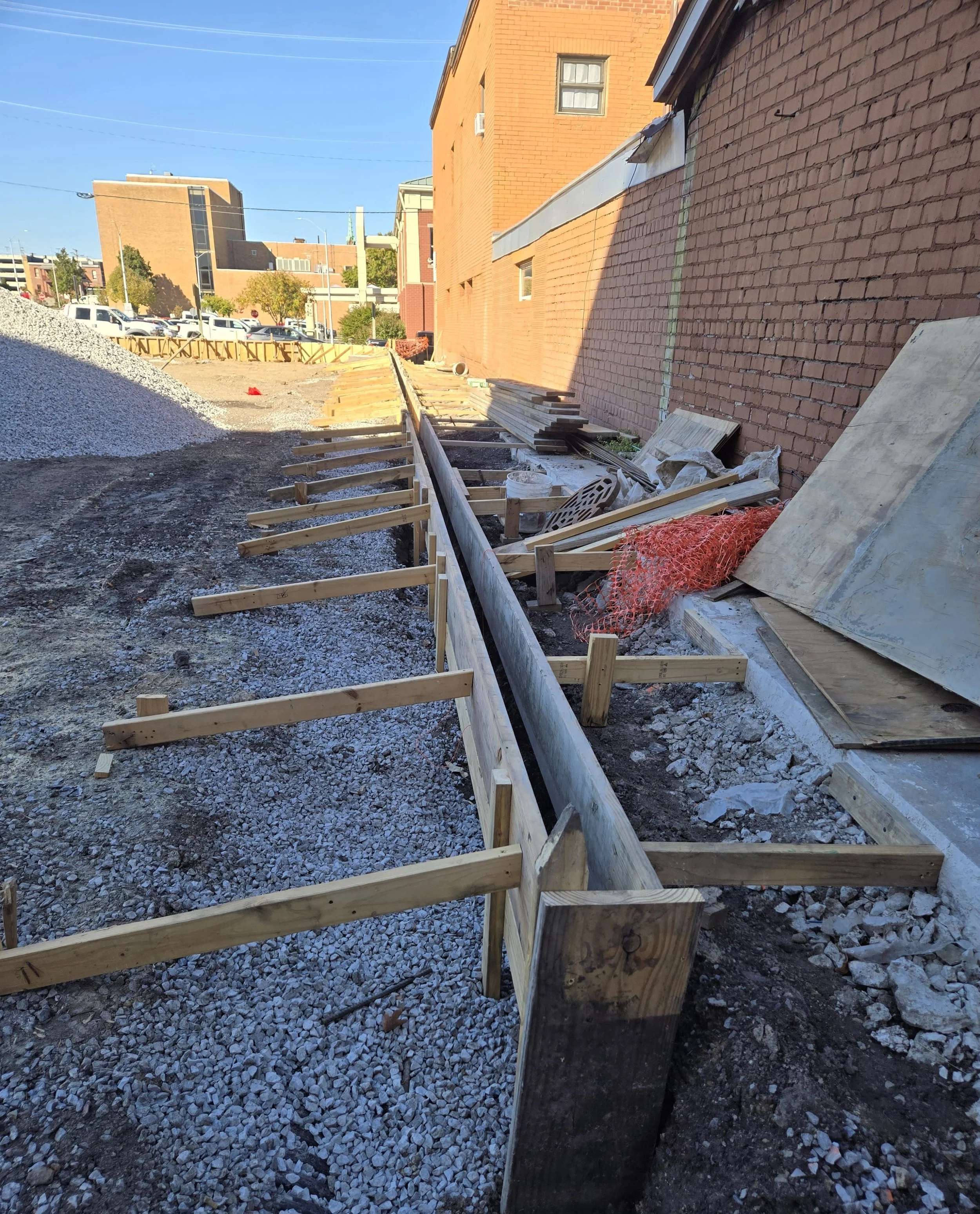Construction site with wooden formwork along a sidewalk next to a brick building, with construction materials and gravel on the ground.