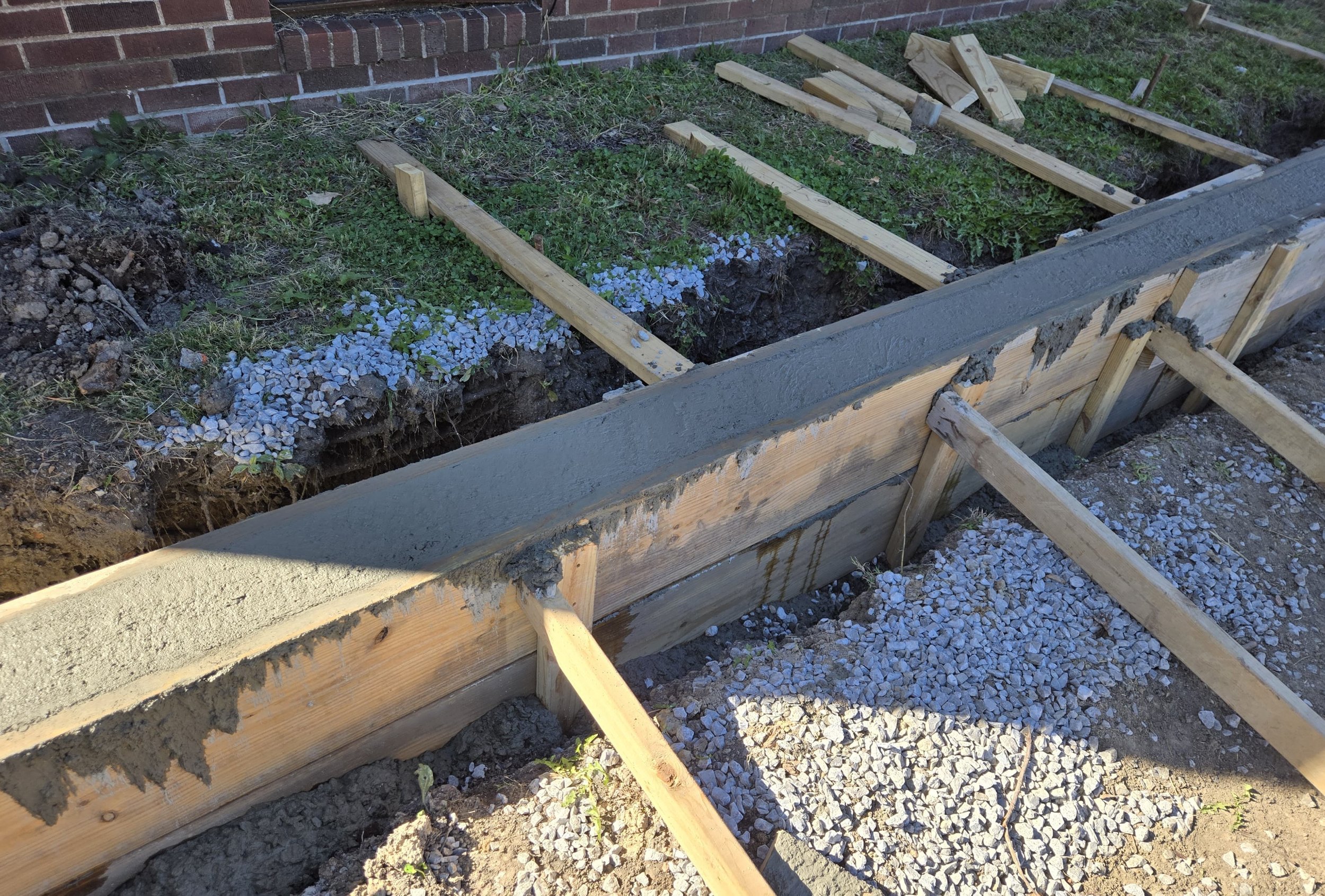 Construction of a concrete curb with wooden supports and gravel foundation, next to a brick wall and grass.