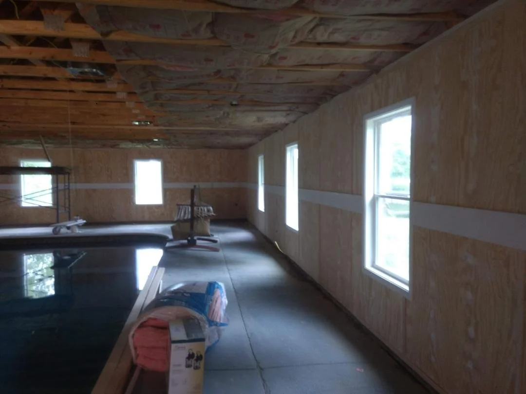 Interior of a room under construction with insulation on the ceiling, plywood walls, and a concrete floor. There are four windows along one wall, and various construction materials and tools are on the floor, including a rolled-up pink insulation bla