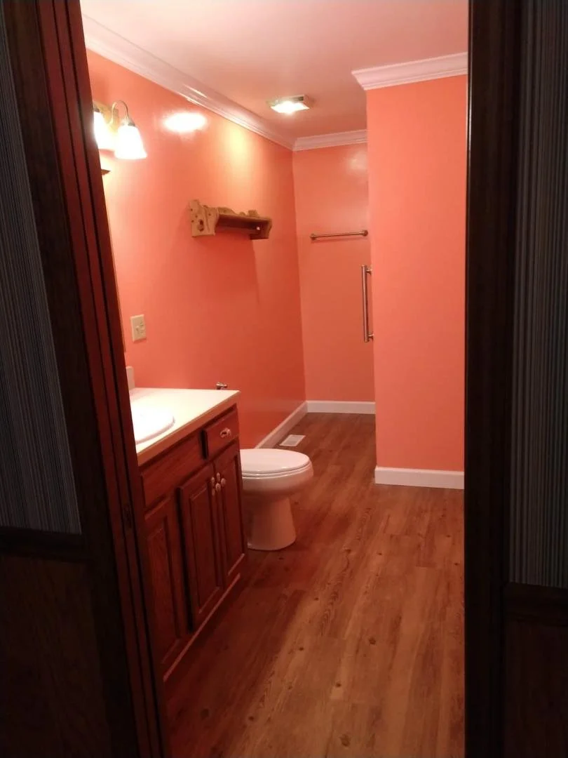 View into a small bathroom with peach-colored walls, wooden cabinet vanity, toilet, and hardwood floor, with a mirror and light fixtures above the sink.
