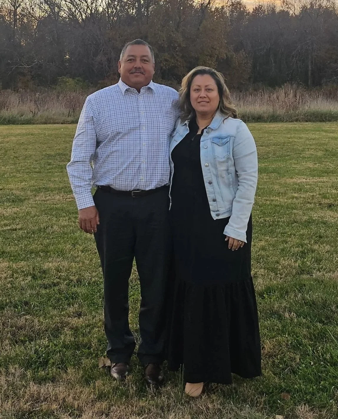 A man and woman standing outdoors on a grassy field with trees in the background, during sunset or late afternoon.