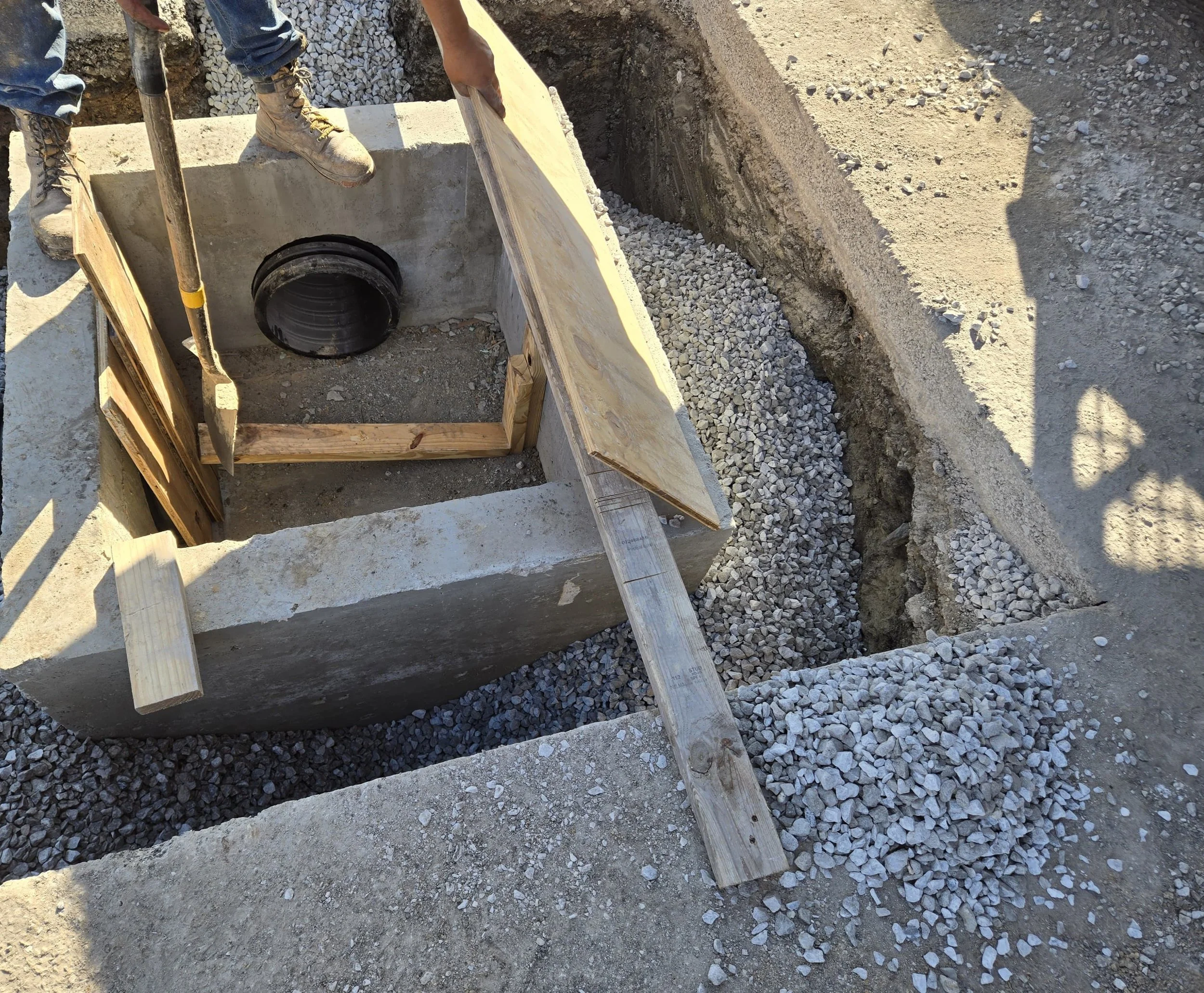 Construction site showing a concrete structure with a pipe fitting; workers are installing a rectangular concrete form with wooden supports and gravel surrounding the site.