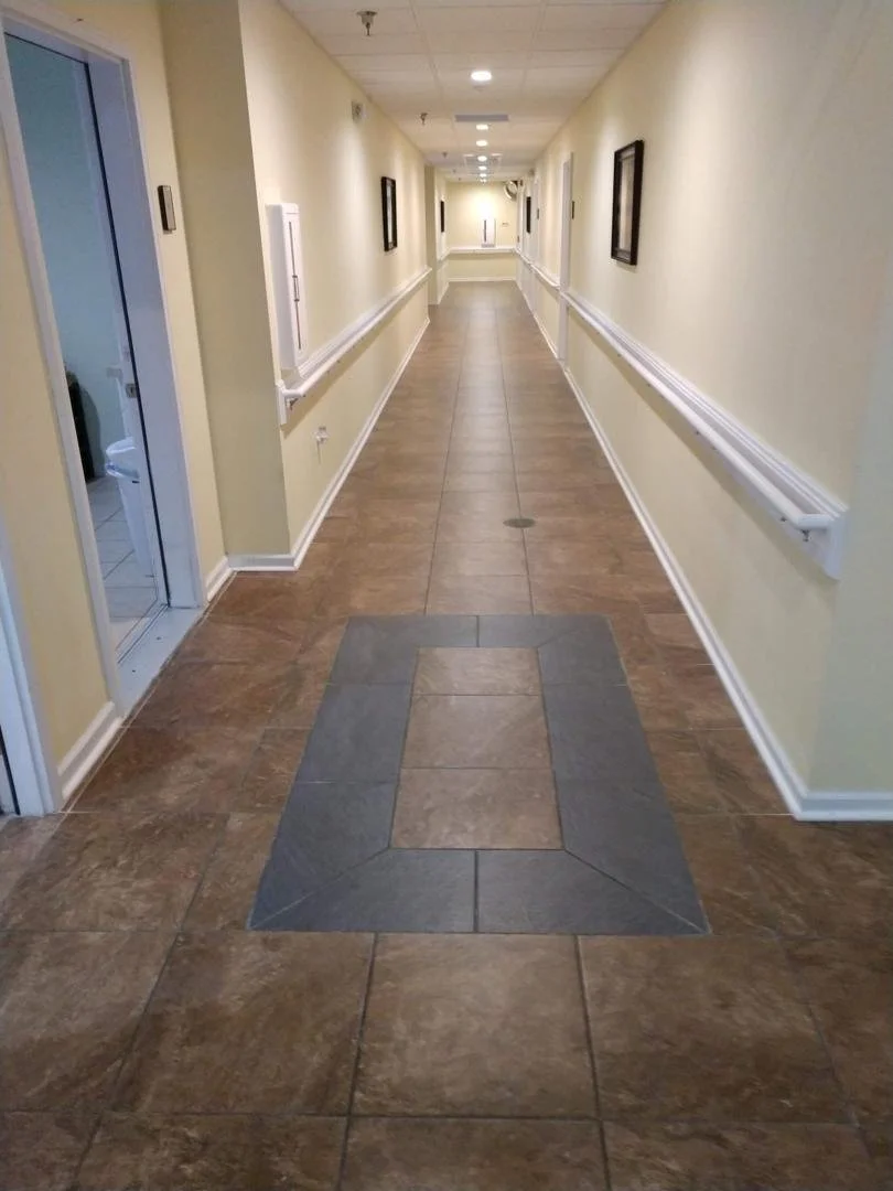 Long indoor hallway with beige walls, framed pictures, and a brown tiled floor with a gray tile border pattern.