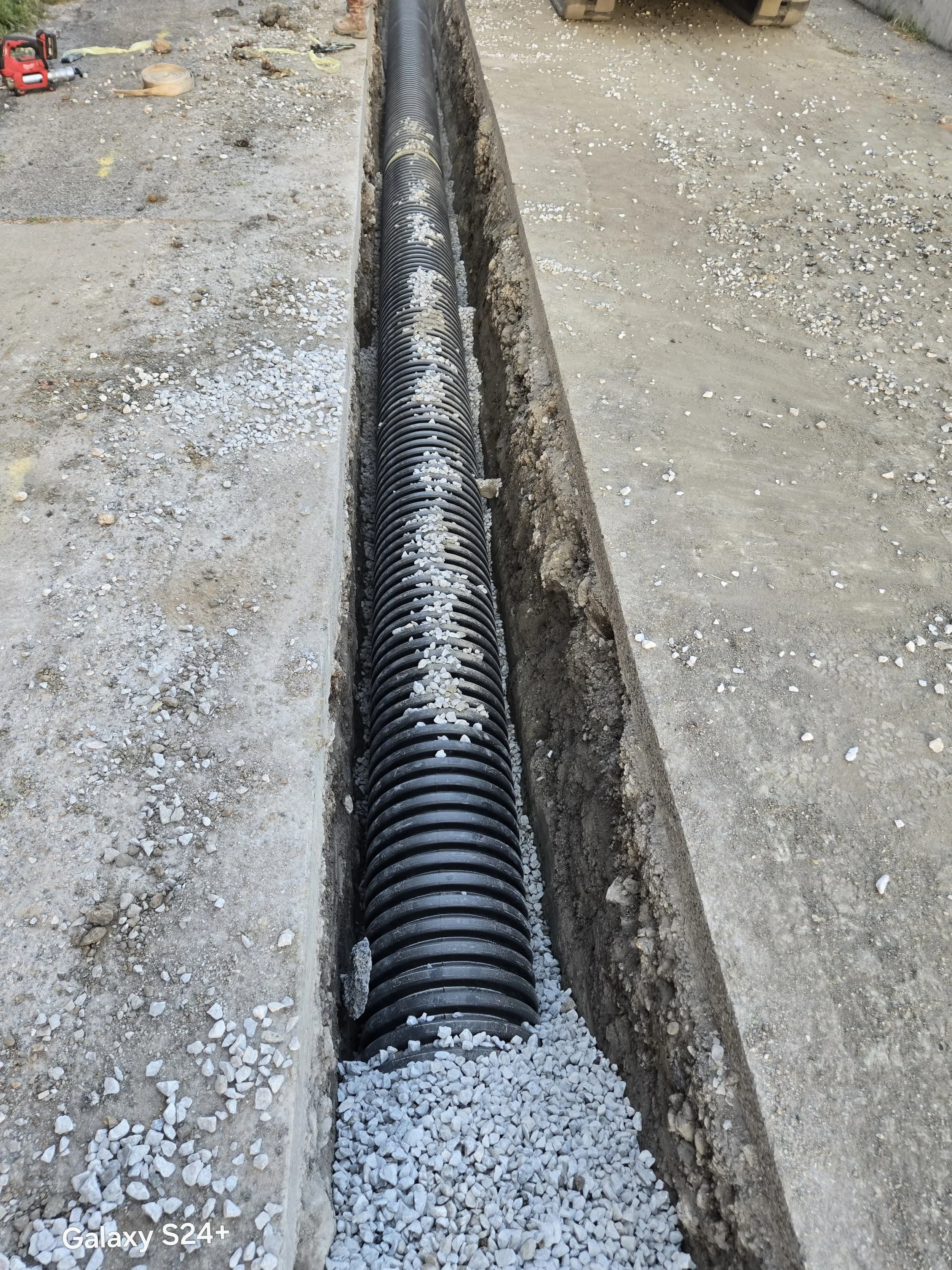 A trench with black corrugated pipe for drainage or utility installation being laid underground at a construction site, with gravel at one end of the pipe.