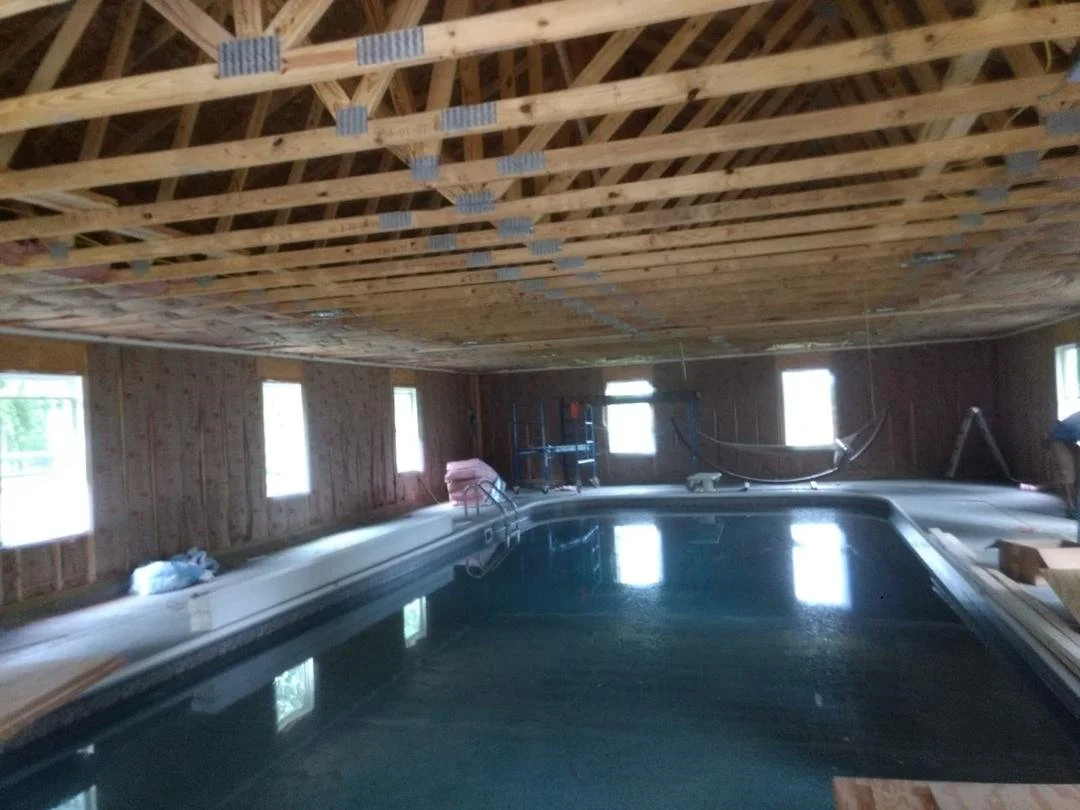 Indoor swimming pool under construction with exposed wooden ceiling and walls, multiple windows, and construction tools around.