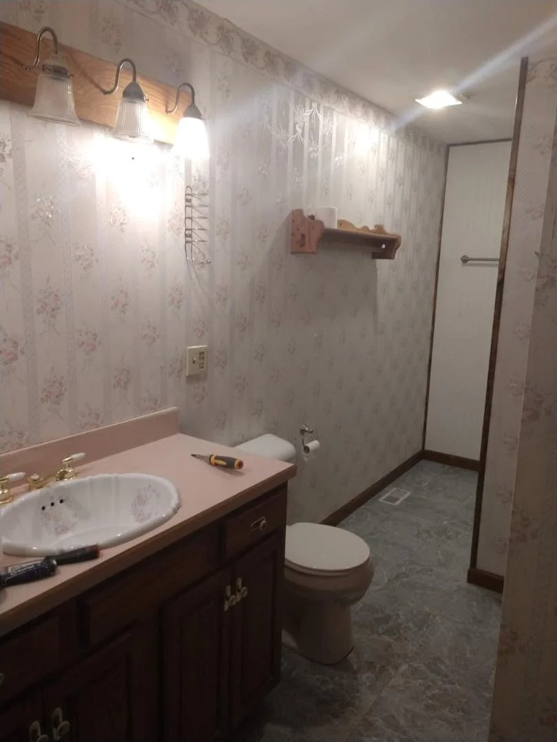 A bathroom with a floral patterned wallpaper, a pink countertop with a white sink, a wooden cabinet below, and a toilet next to a wall with a small wooden shelf. There is a mirror on the wall, a towel rack in the shower area, and construction tools o
