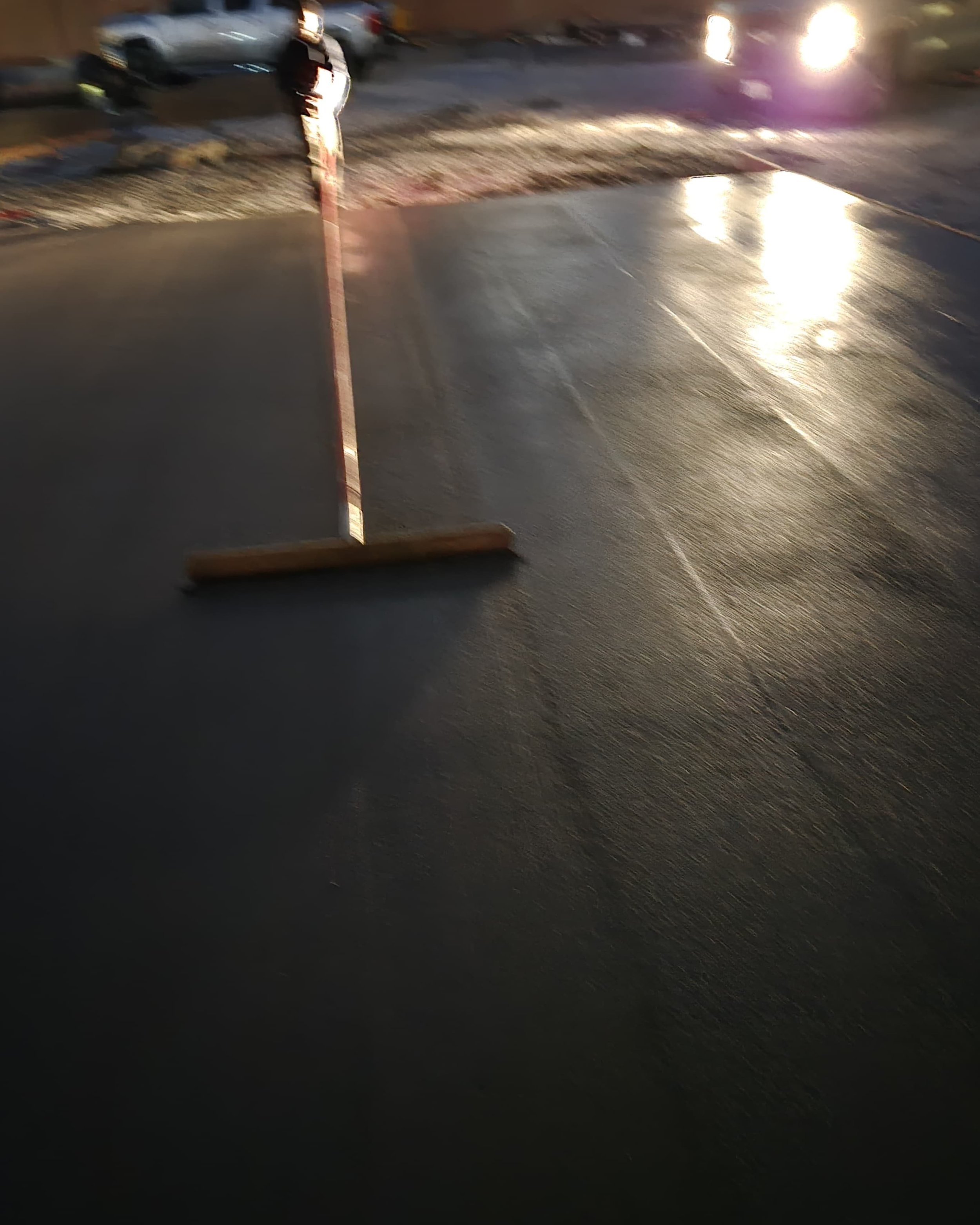 A person working on a freshly paved blacktop surface with a roller tool, near parked cars in a parking lot during evening or night.