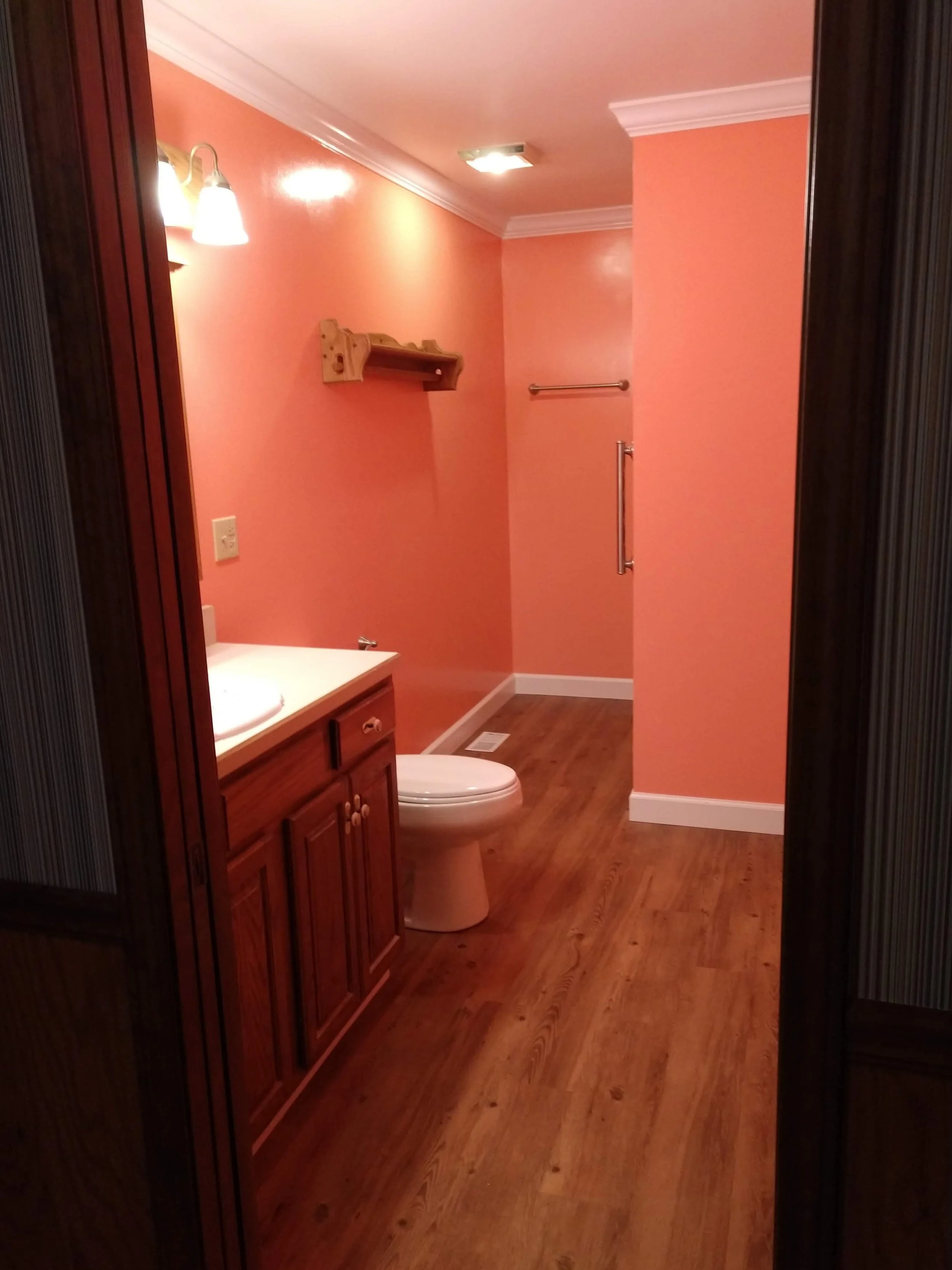 Photo of a bathroom with pink walls, wooden cabinetry, a toilet, and hardwood flooring, with a mirror reflecting part of the room.